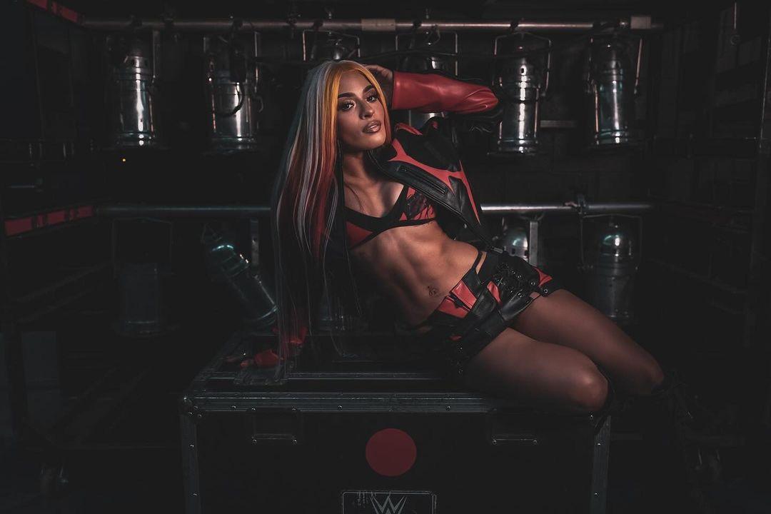 Zelina Vega in a red and black leather jacket, sports bra, and mini skirt posing in a dark industrial setting