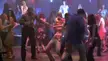 A shirtless man and a group of people in casual club wear dancing in a dimly lit nightclub with blue and red lighting