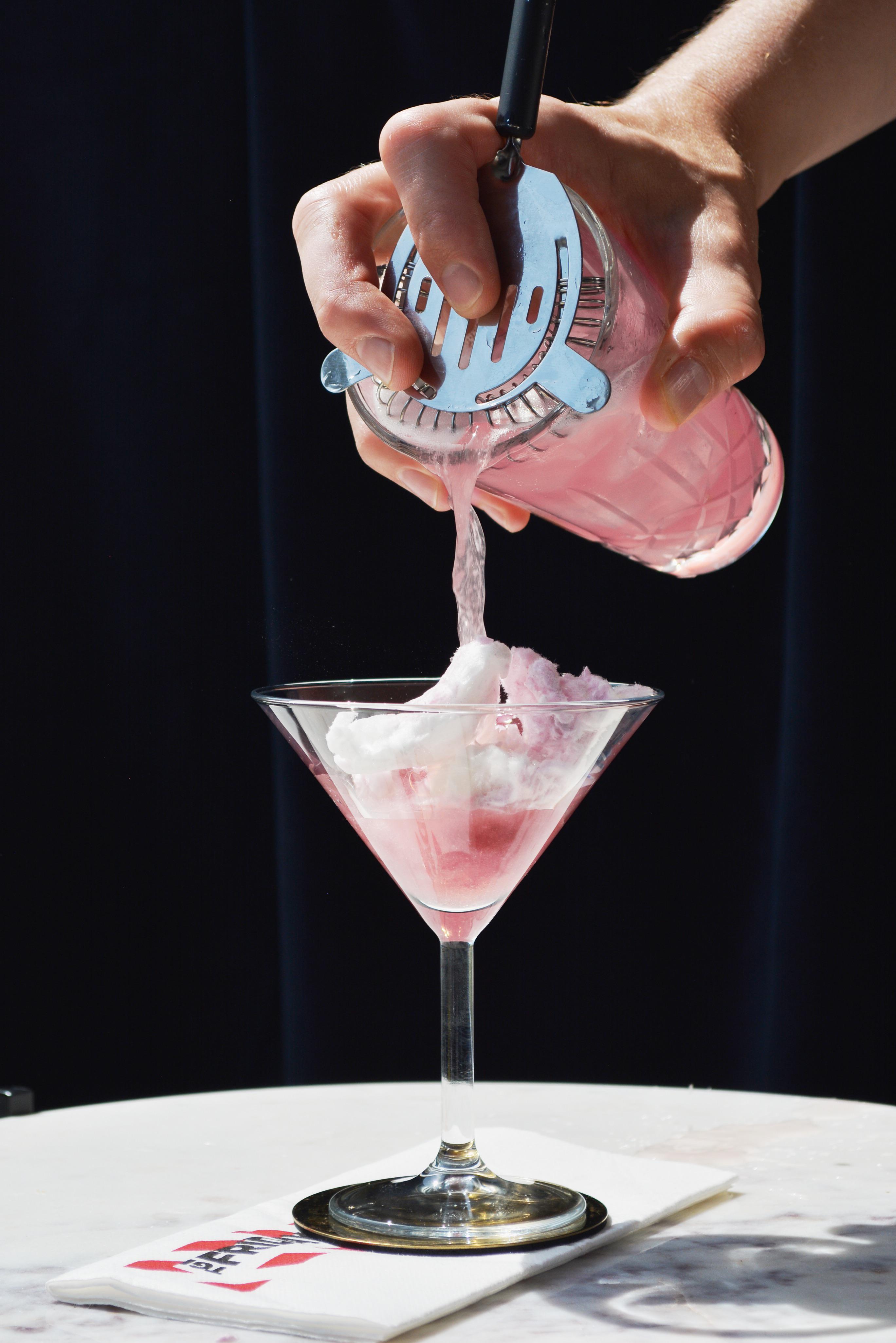 A person with pale skin pours a pink Punk Cosmo cocktail from a shaker into a martini glass with white foam.