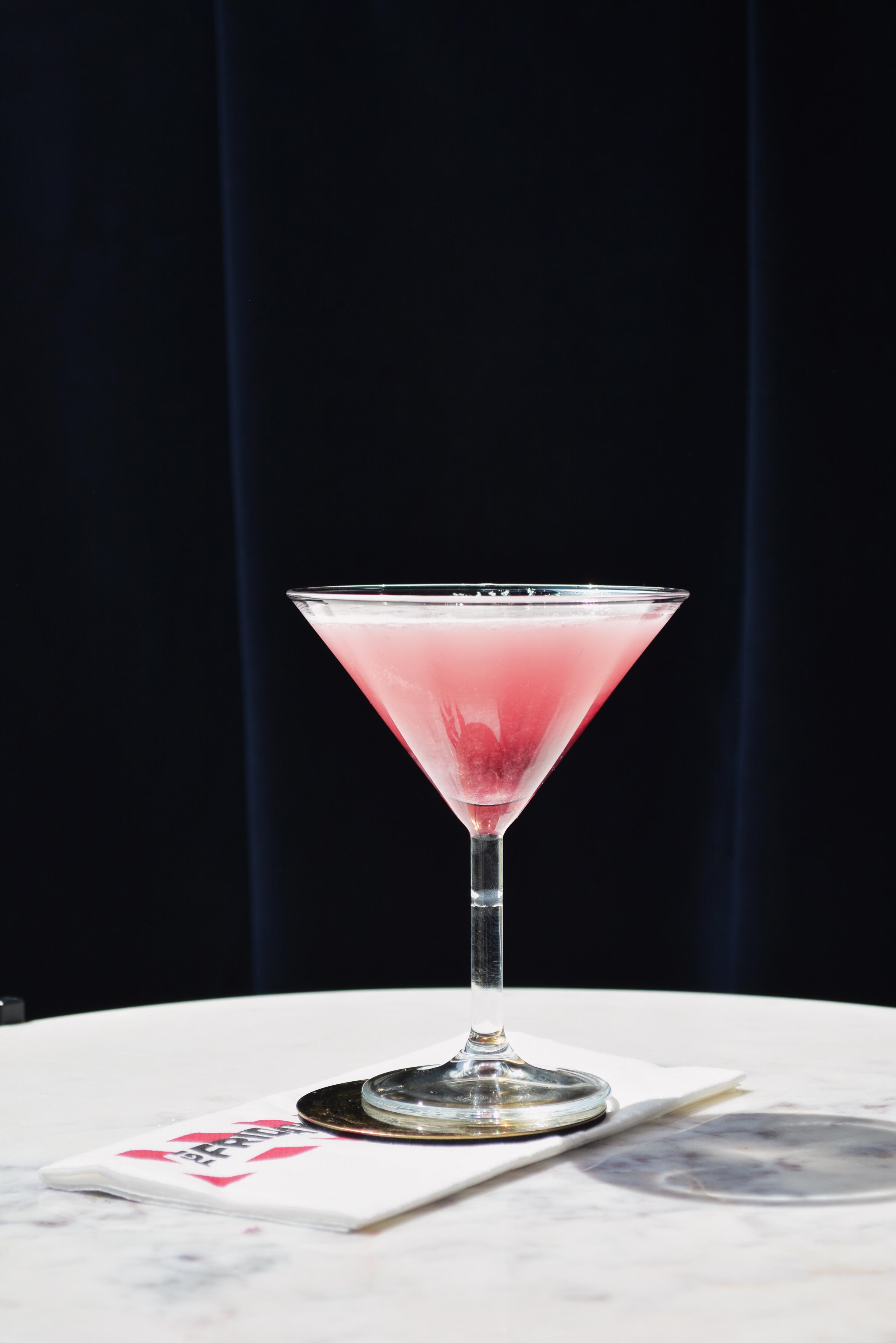 A person with pale skin pours a pink Punk Cosmo cocktail from a shaker into a martini glass with white foam.