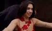 Dua Lipa in a red floral dress having a leather bracer strapped to her arm by a man in a suit