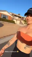 Athletic white woman with tattoos and a nose piercing in an orange tube top and black Nike cap posing for a selfie video