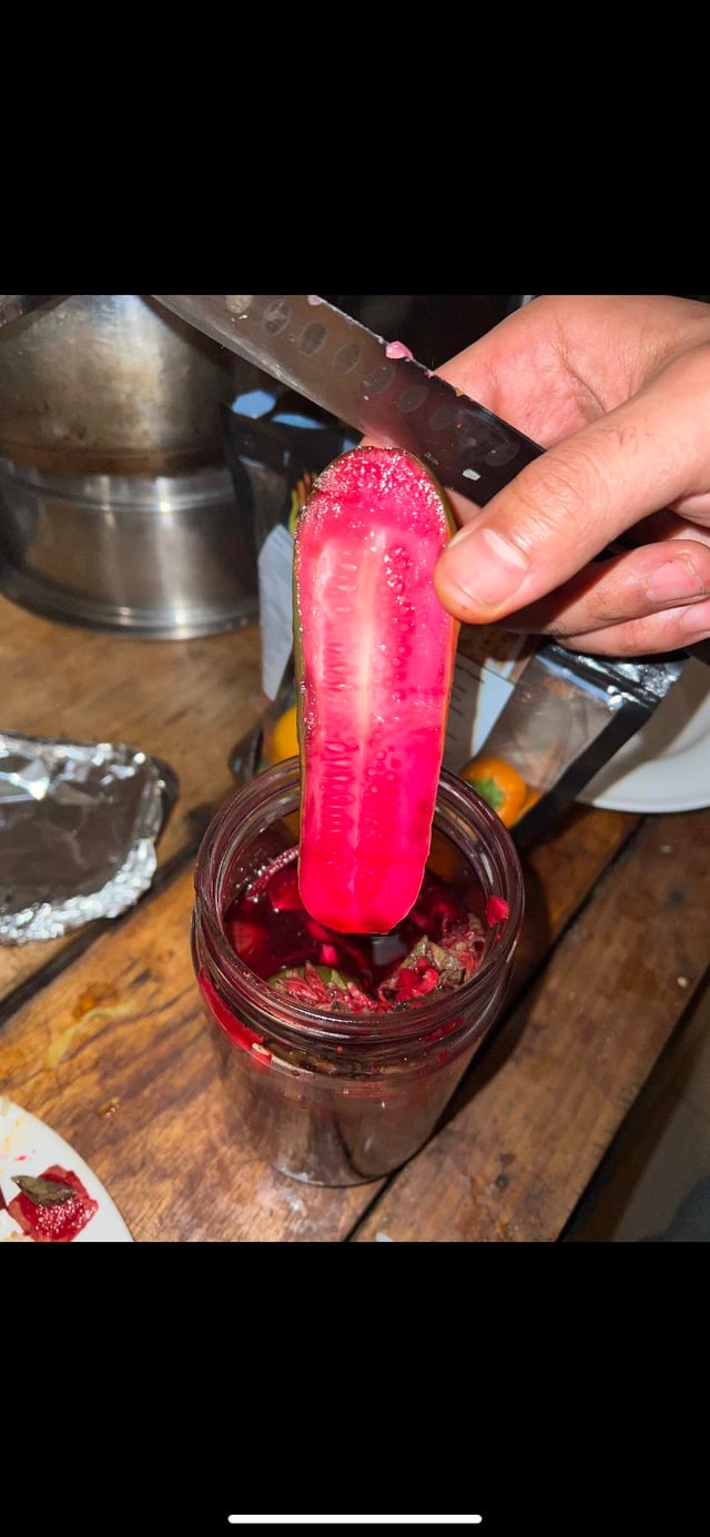 A person with pale skin holds a slice of pink pickle over a glass jar on a wooden table.