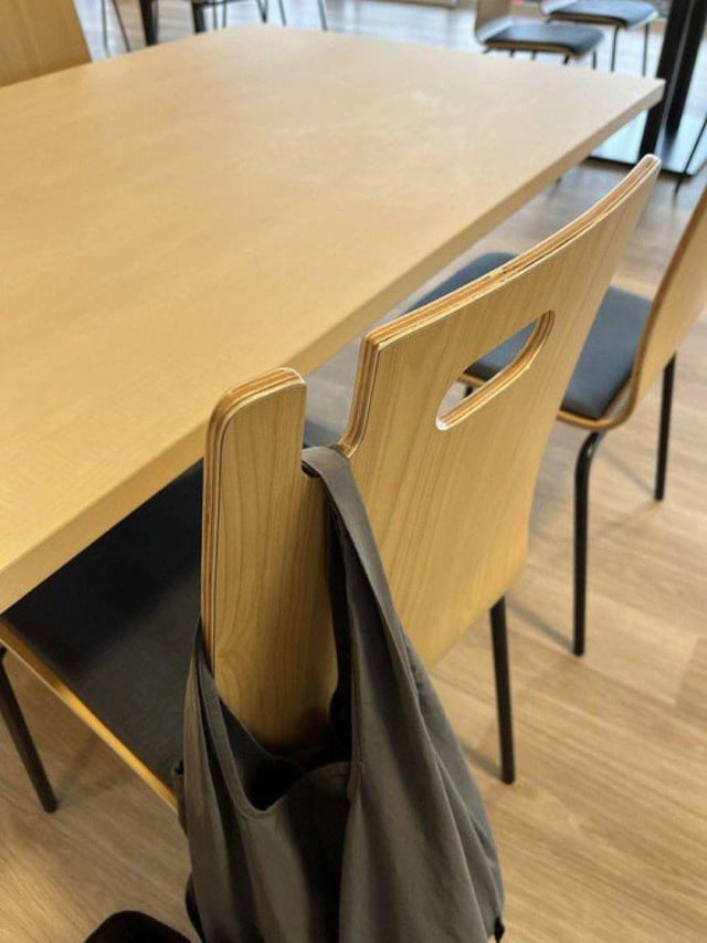 A black bag hanging from a light-colored wooden chair next to a matching table in a casual interior design setting.