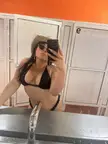 A curvy trans woman in a black bikini, straw hat, and sunglasses taking a mirror selfie in a bathroom.
