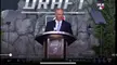 A man in a dark suit and blue tie speaking at an MLB Draft podium with a white shirt.