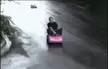 A man in a black t-shirt riding a small pink toy car on a wet road, crashing into bushes and flipping over.