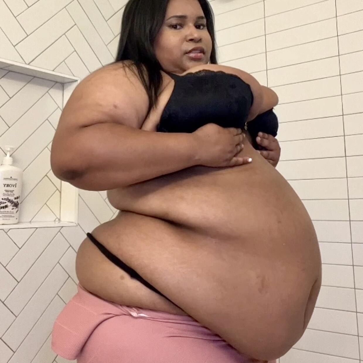 BigBellyJudy, a beautiful ebony bbw, posing in a black lace bra and pink pants in a bathroom selfie