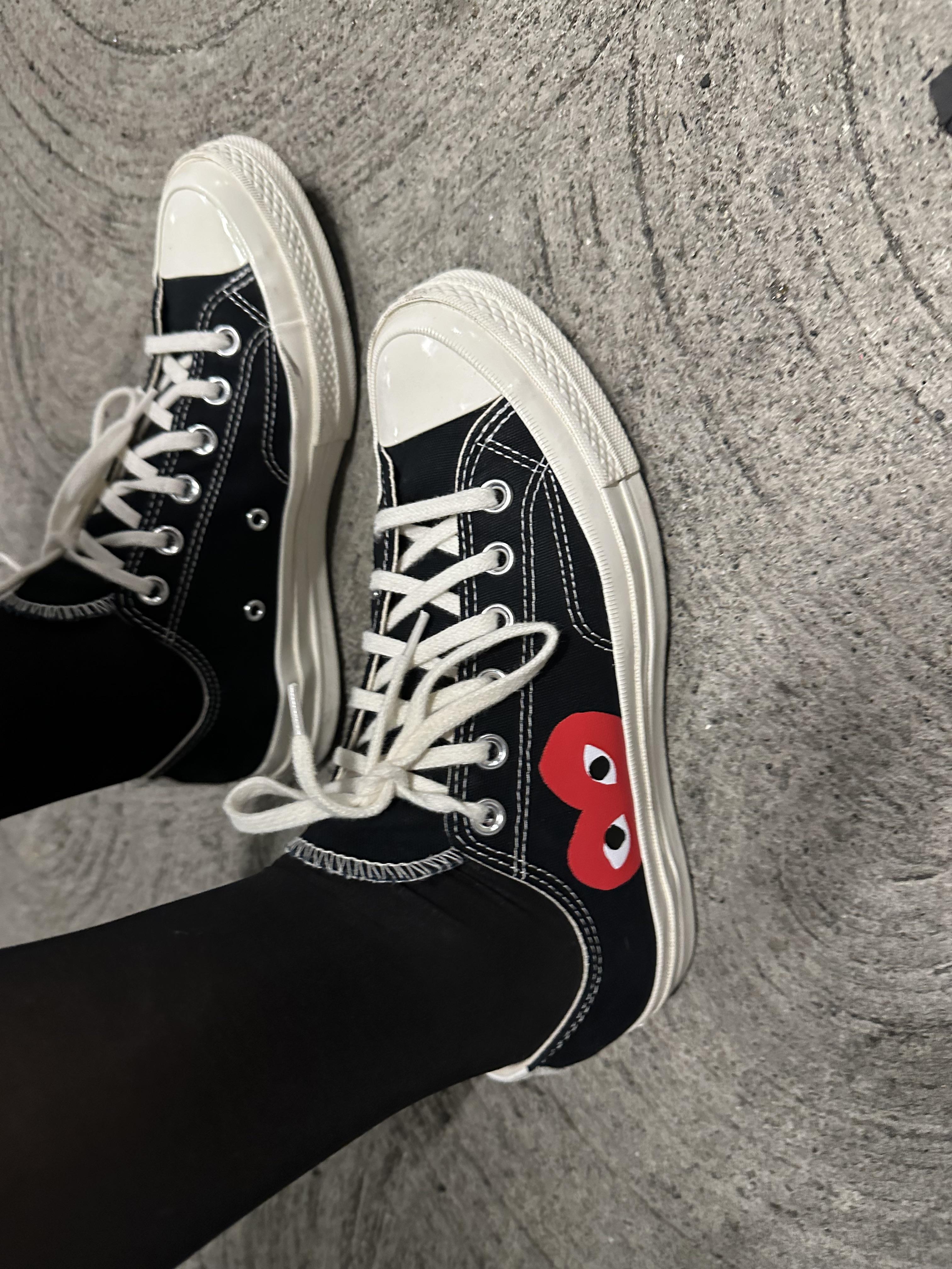 Top-down view of black Converse sneakers with white laces and a red eye logo, worn with black tights on a concrete floor.