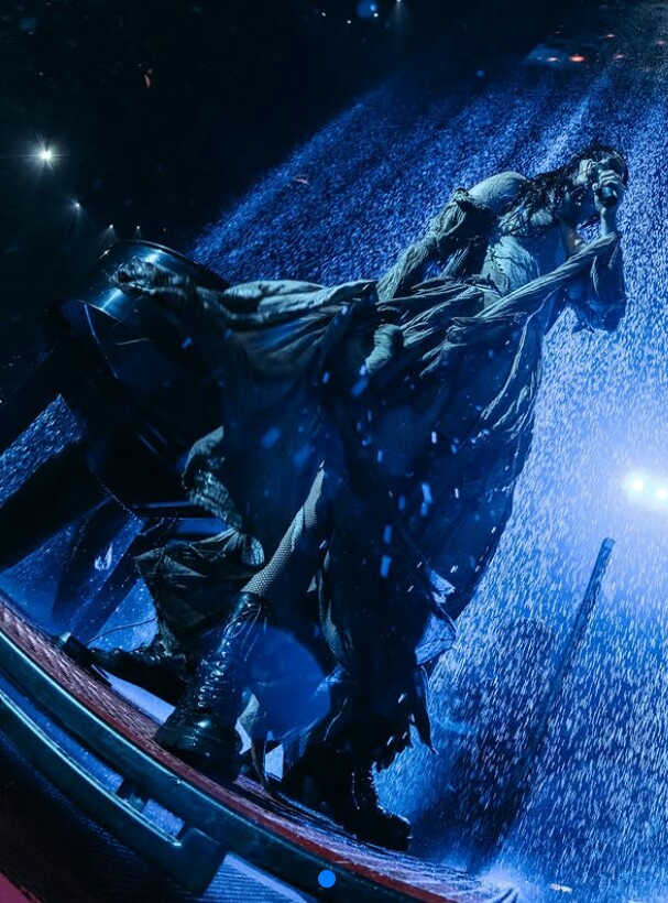 Davina Michelle in a tattered fantasy dress and corset, singing under rain with wet black hair and a slim build.