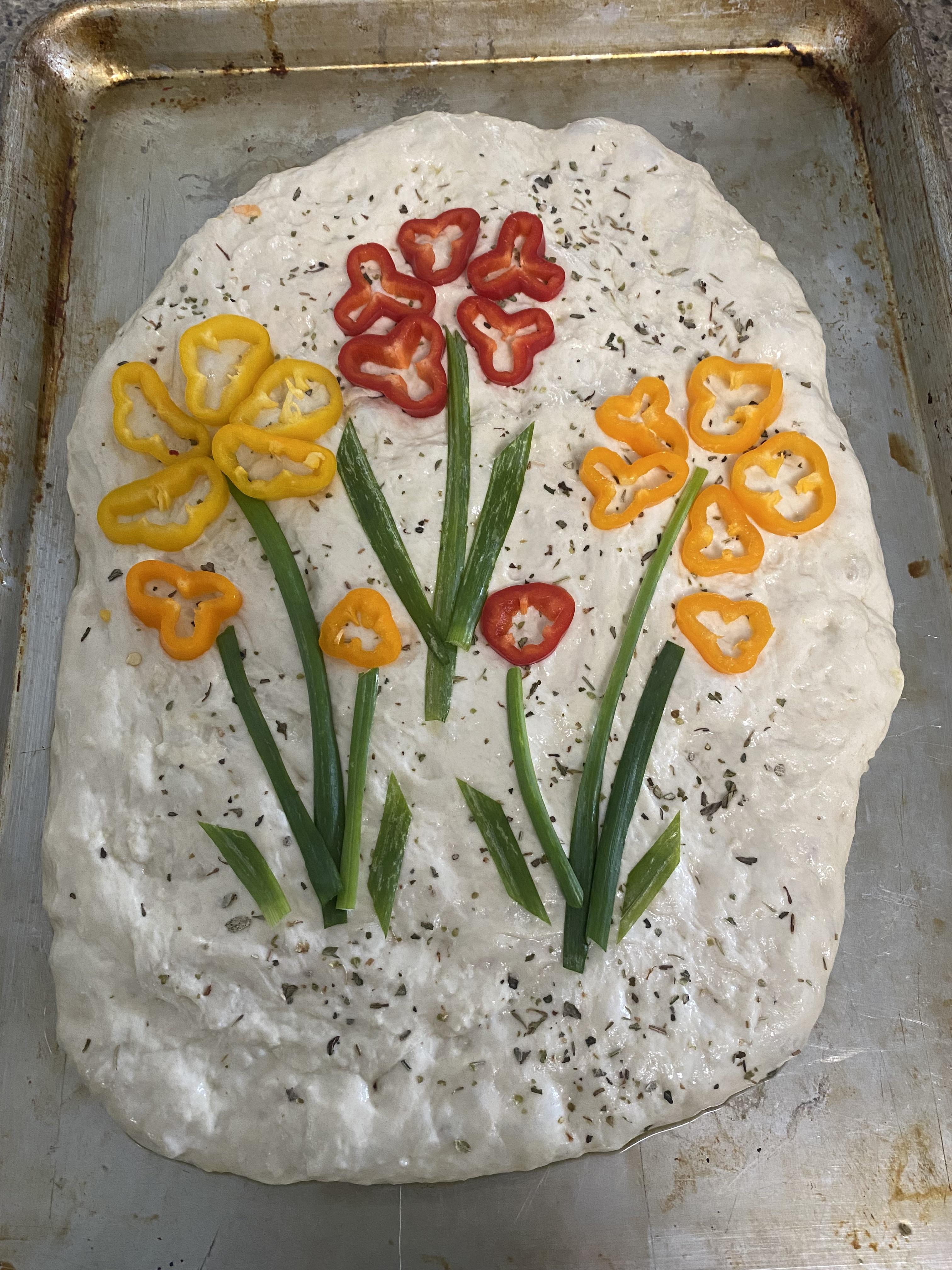 Homemade focaccia flatbread decorated with bell pepper flowers on a baking sheet before and after baking