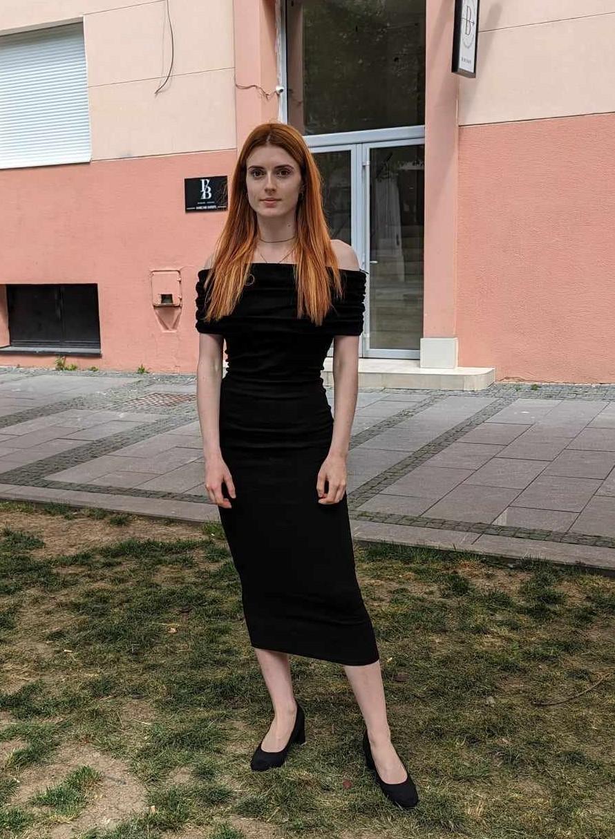 A slender woman with long red hair and pale skin wearing a black t-shirt, blue tank top, and black midi dress.