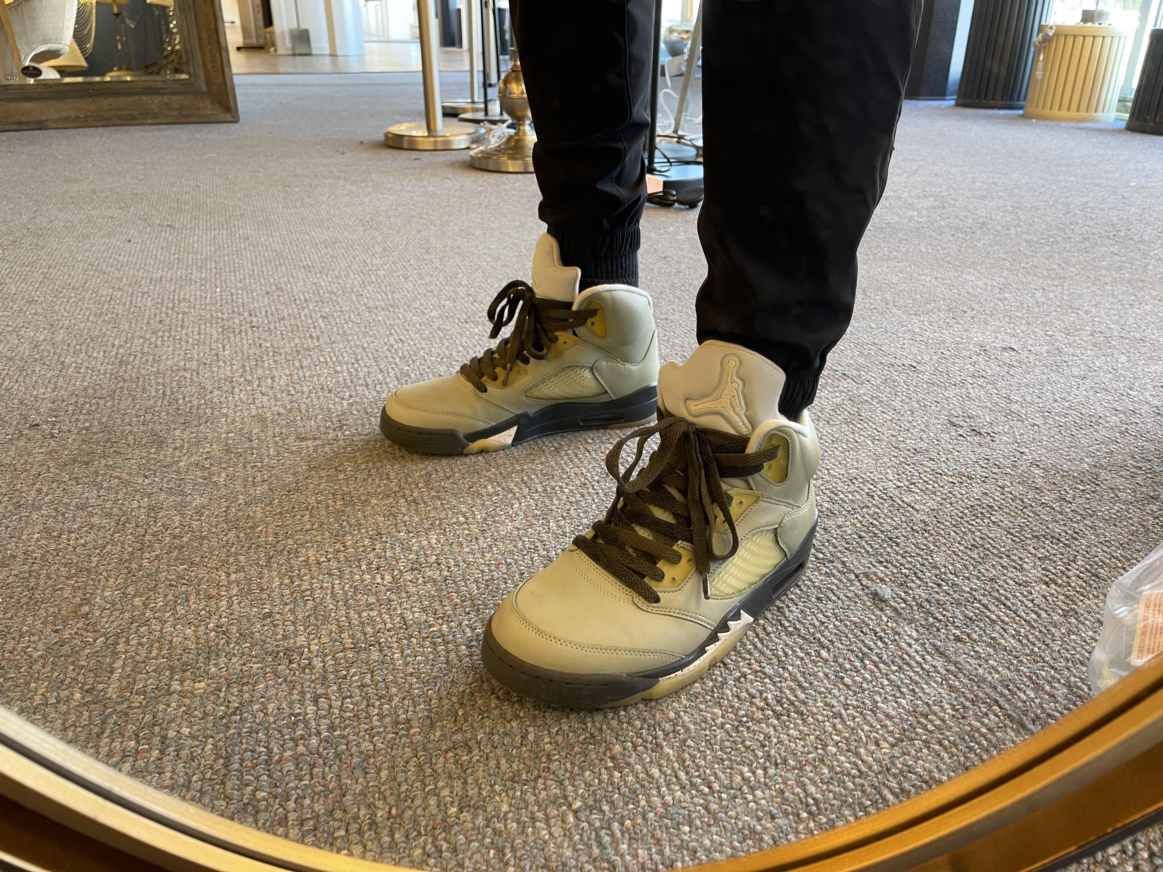 A person in black pants and light-colored high-top sneakers on a grey carpet, showcasing a lace swap.