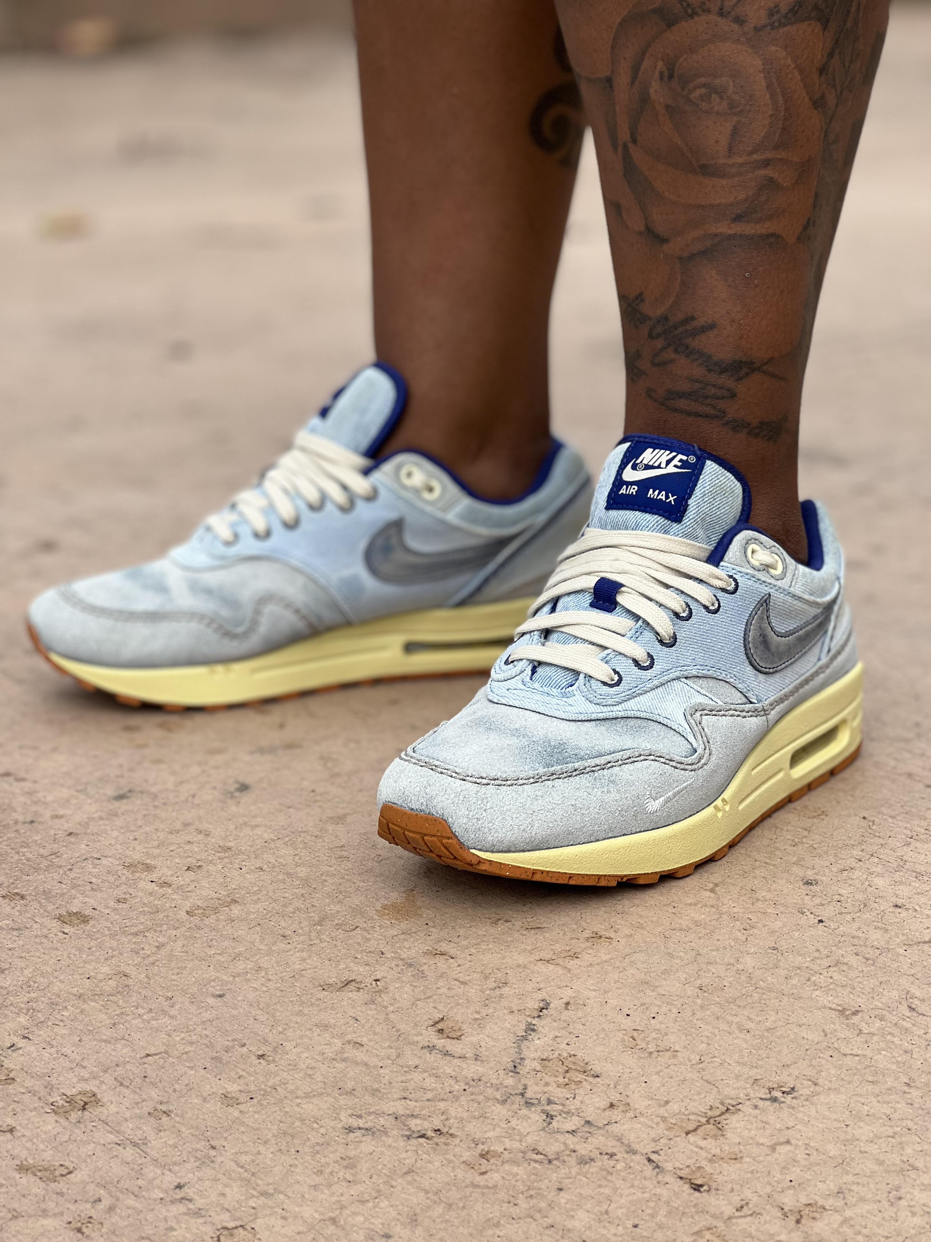 Close-up of light blue Nike Air Max 1 Dirty Denim sneakers on a person with dark skin and a floral leg tattoo.