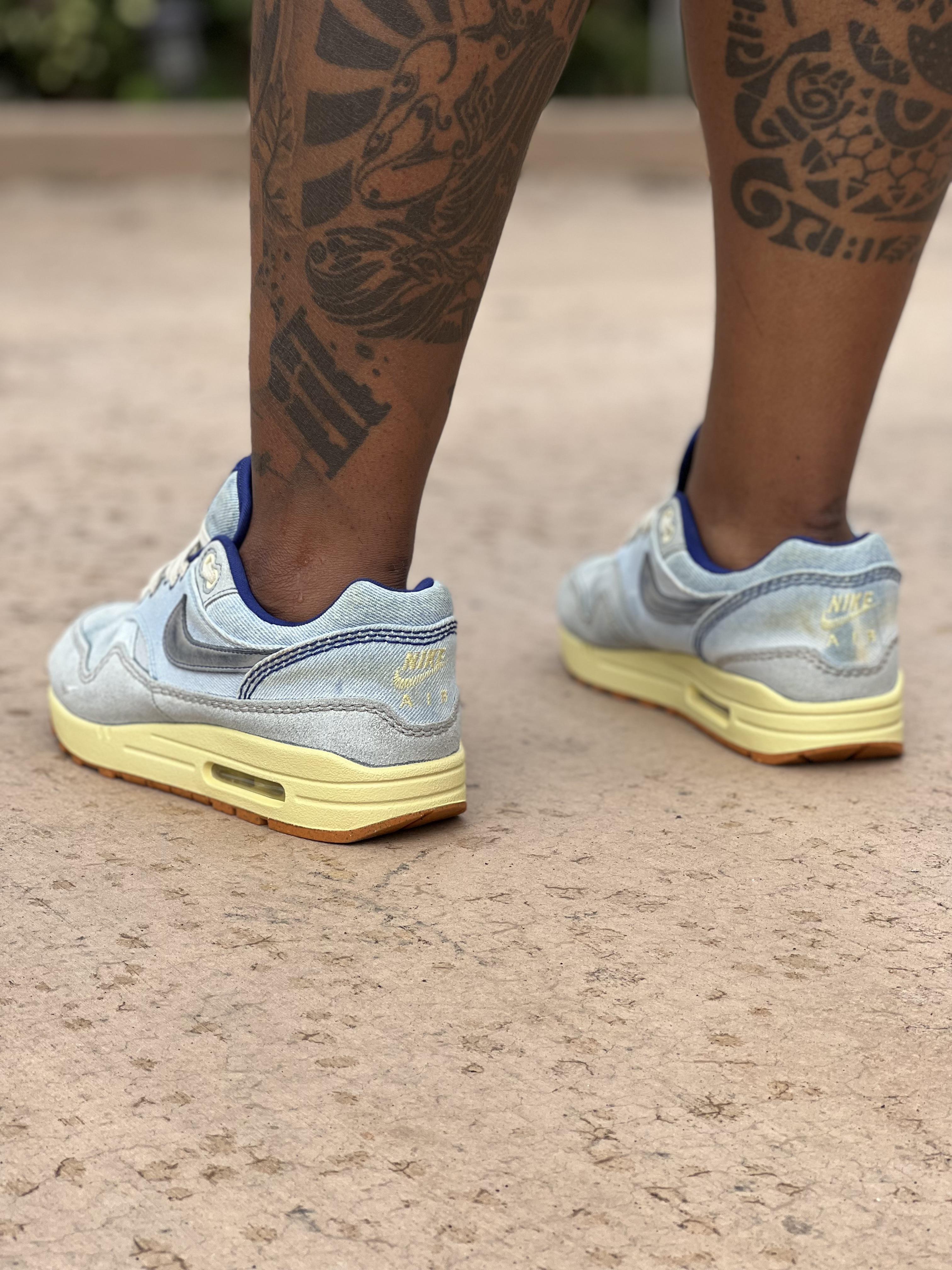 Close-up of light blue Nike Air Max 1 Dirty Denim sneakers on a person with dark skin and a floral leg tattoo.