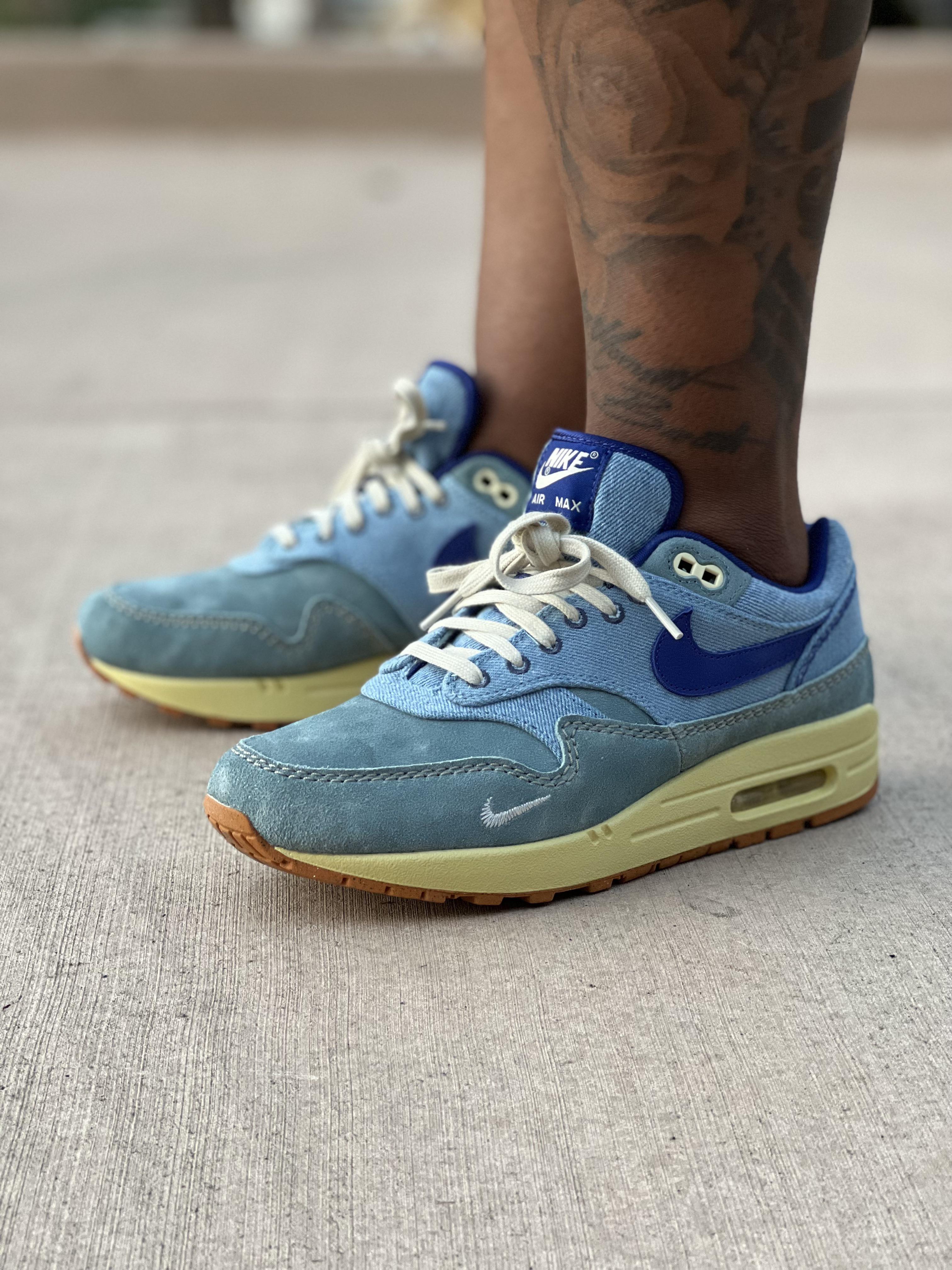 Close-up of light blue Nike Air Max 1 Dirty Denim sneakers on a person with dark skin and a floral leg tattoo.