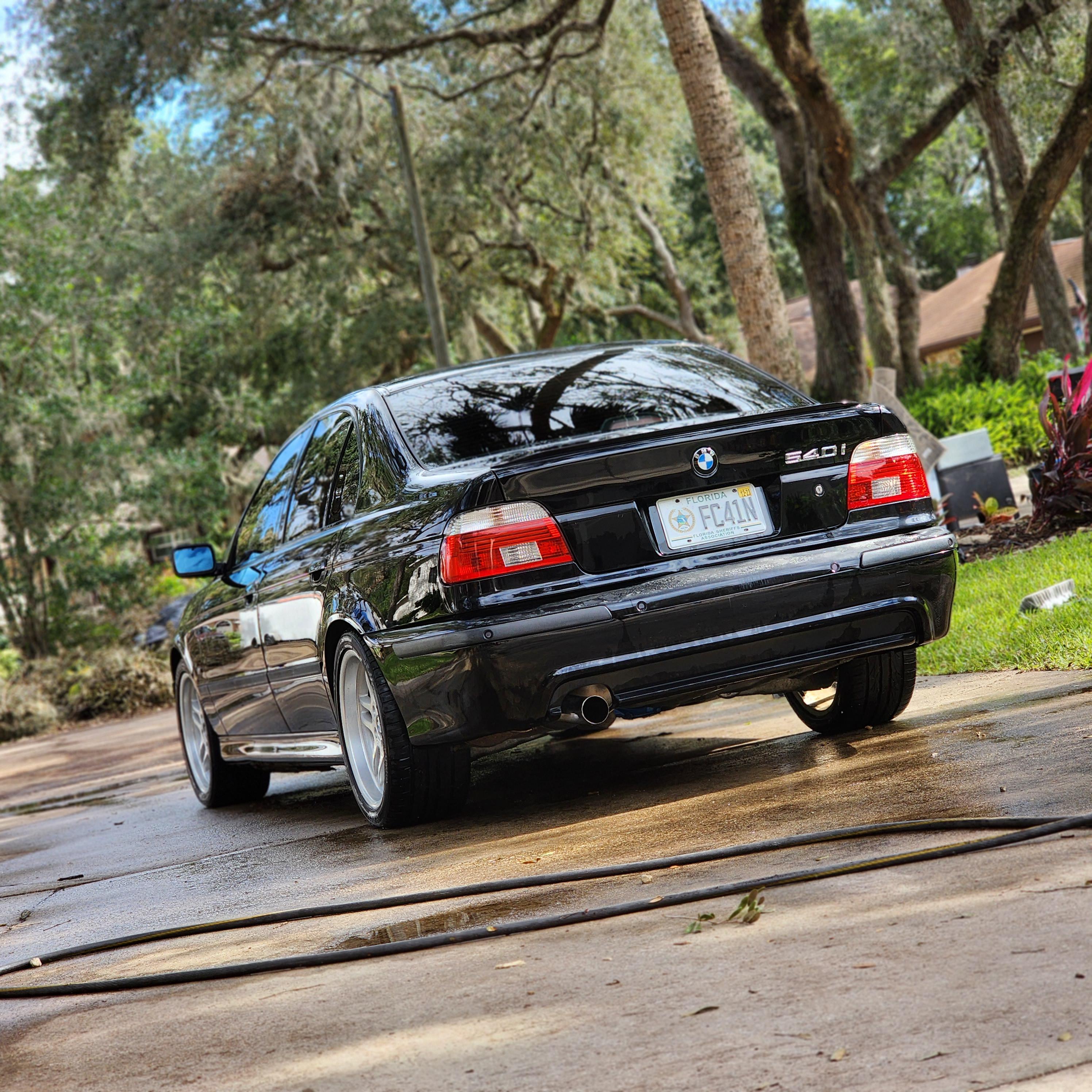 Back view of a black BMW 540i sedan parked on a wet concrete driveway surrounded by trees