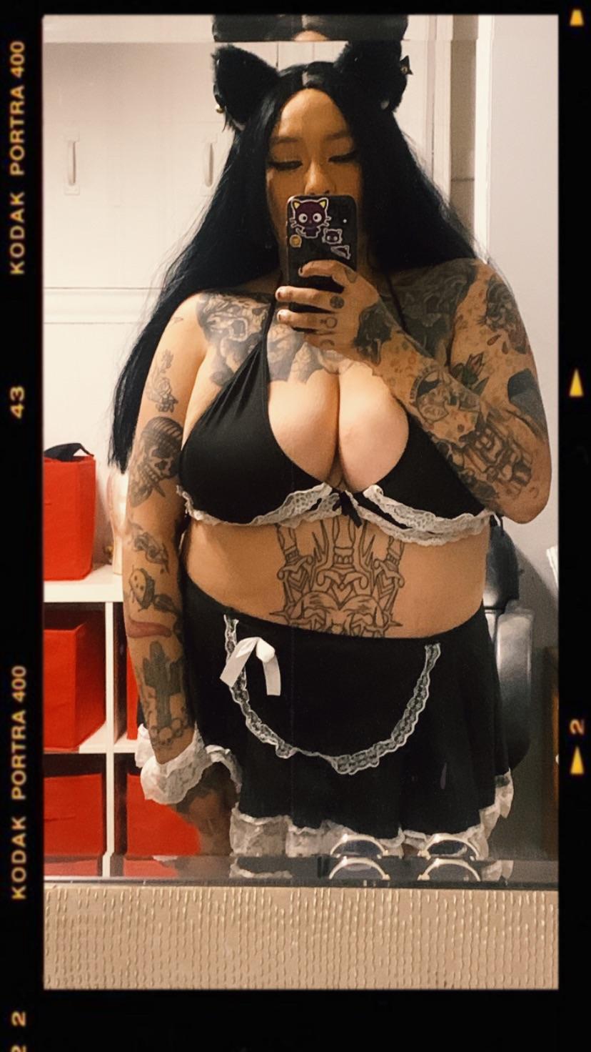 Busty mixed woman with extensive blackout tattoos in a black maid costume posing for selfies