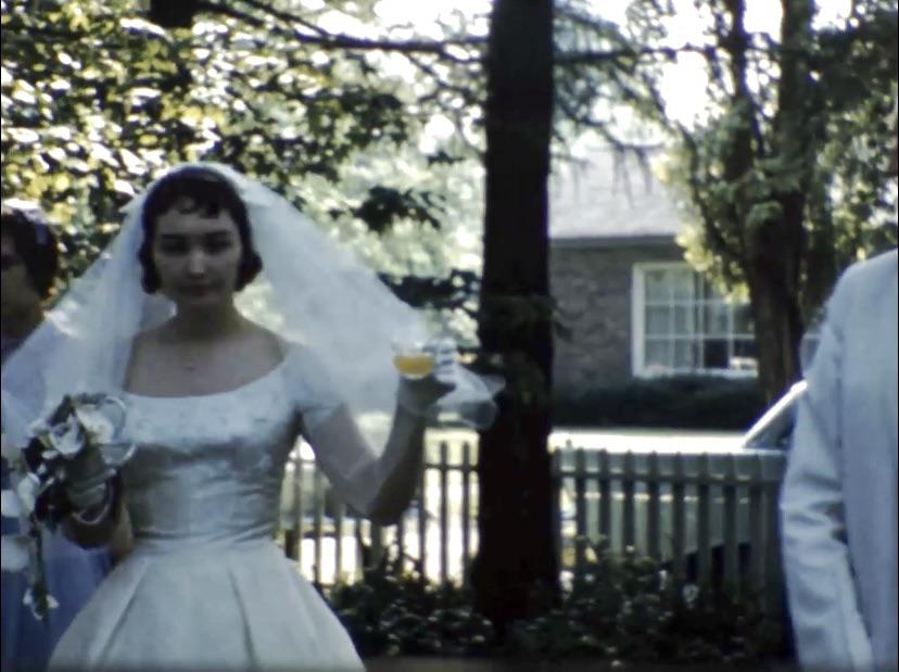 Grandparents in white formal wear and a bridal veil with a wedding party in an outdoor garden by a white fence