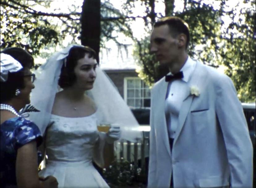Grandparents in white formal wear and a bridal veil with a wedding party in an outdoor garden by a white fence