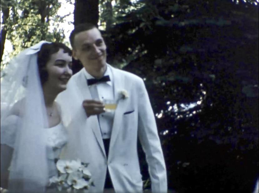 Grandparents in white formal wear and a bridal veil with a wedding party in an outdoor garden by a white fence
