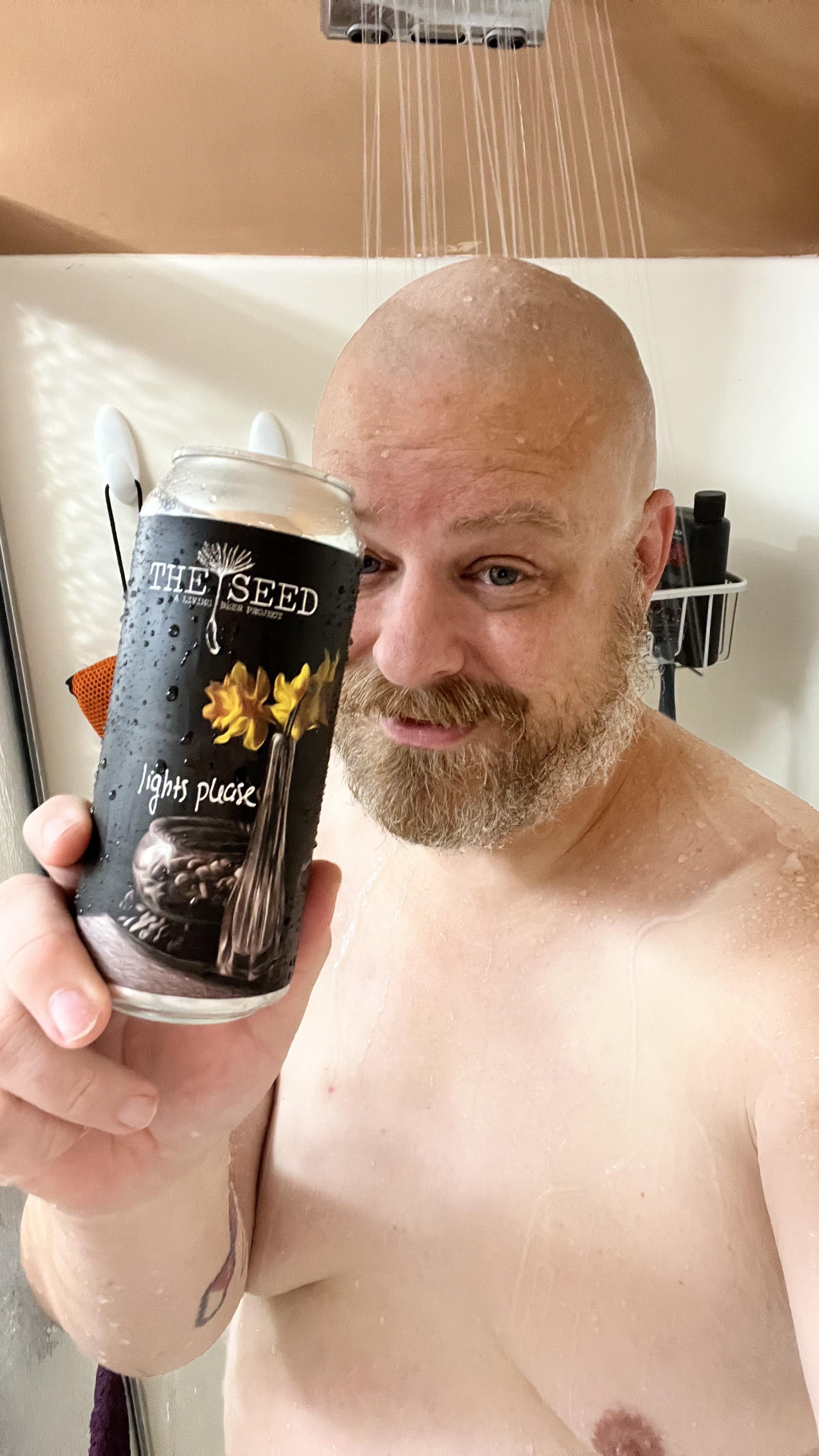 Shirtless man with a beard and arm tattoo holding a Lights Please IPA beer can in a bathroom shower.