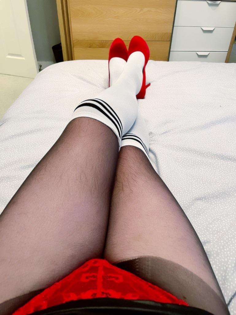 Femboy in a black leather mini skirt, red high heels, and white over-knee socks posing on a bed.