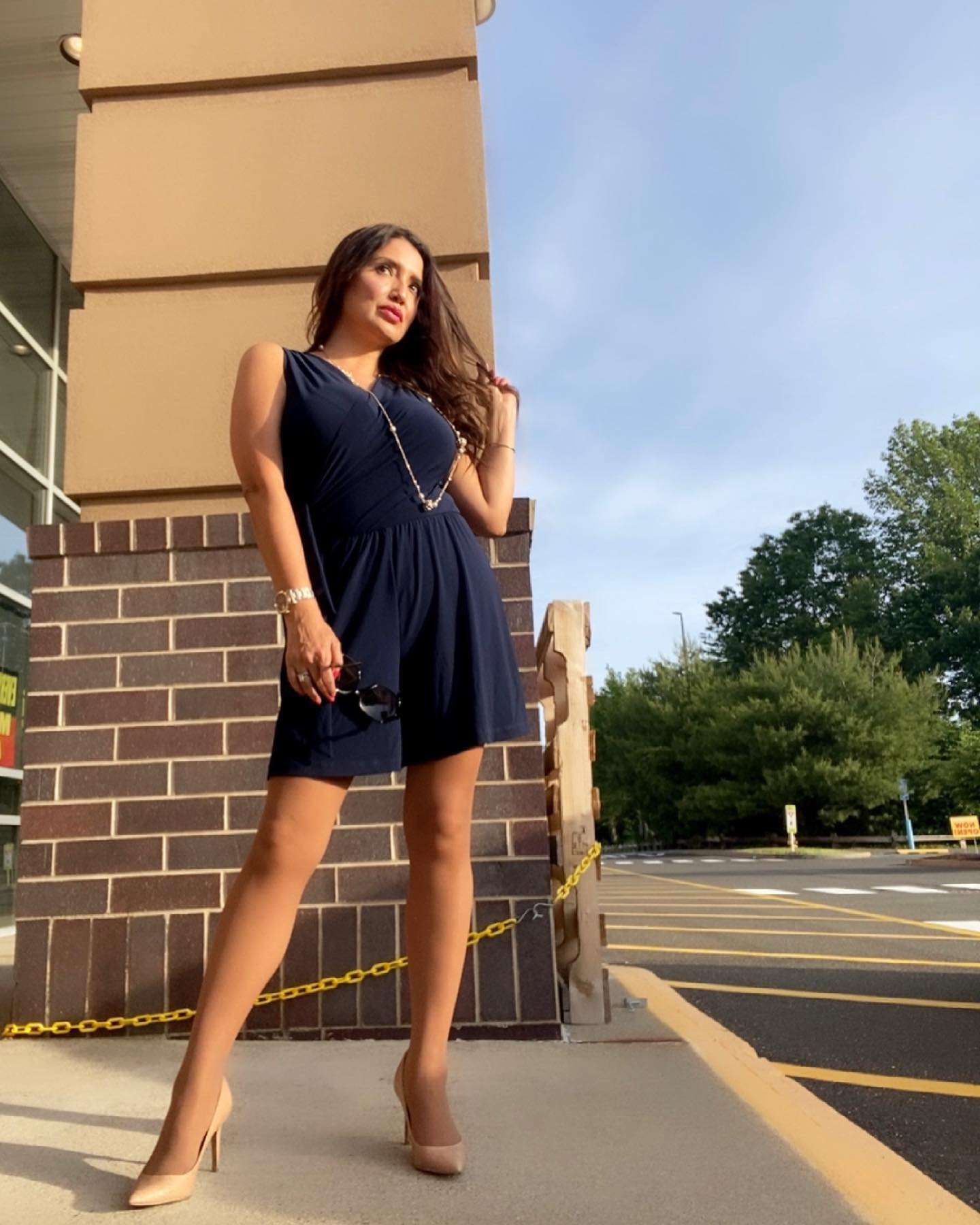 Curvy Indian woman in a navy blue romper and nude heels posing by a brick building