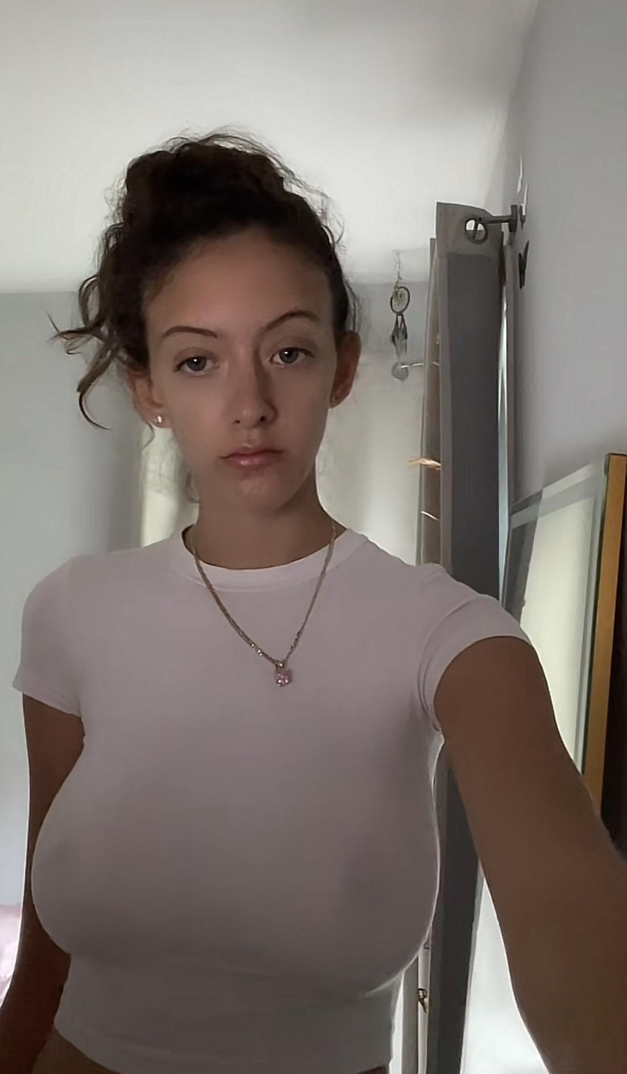 Busty brunette in a tight white t-shirt and grey sweatpants taking a mirror selfie