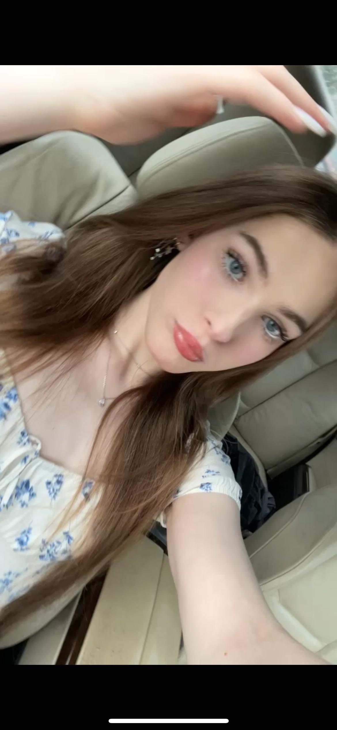 Trans woman with long brown hair and blue eyes in a white floral dress posing for a selfie in a car interior