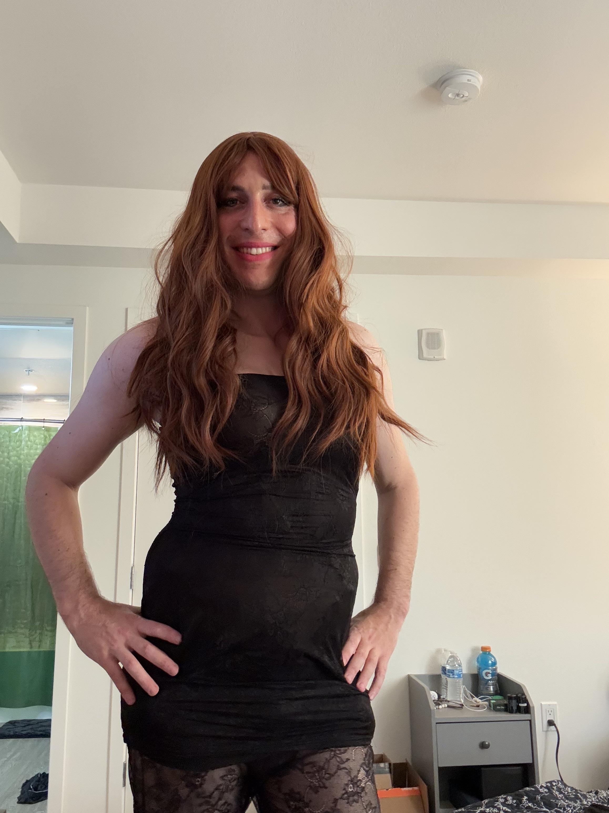 Sissy femboy in a black strapless dress, long red wig, and lace tights posing for a selfie.