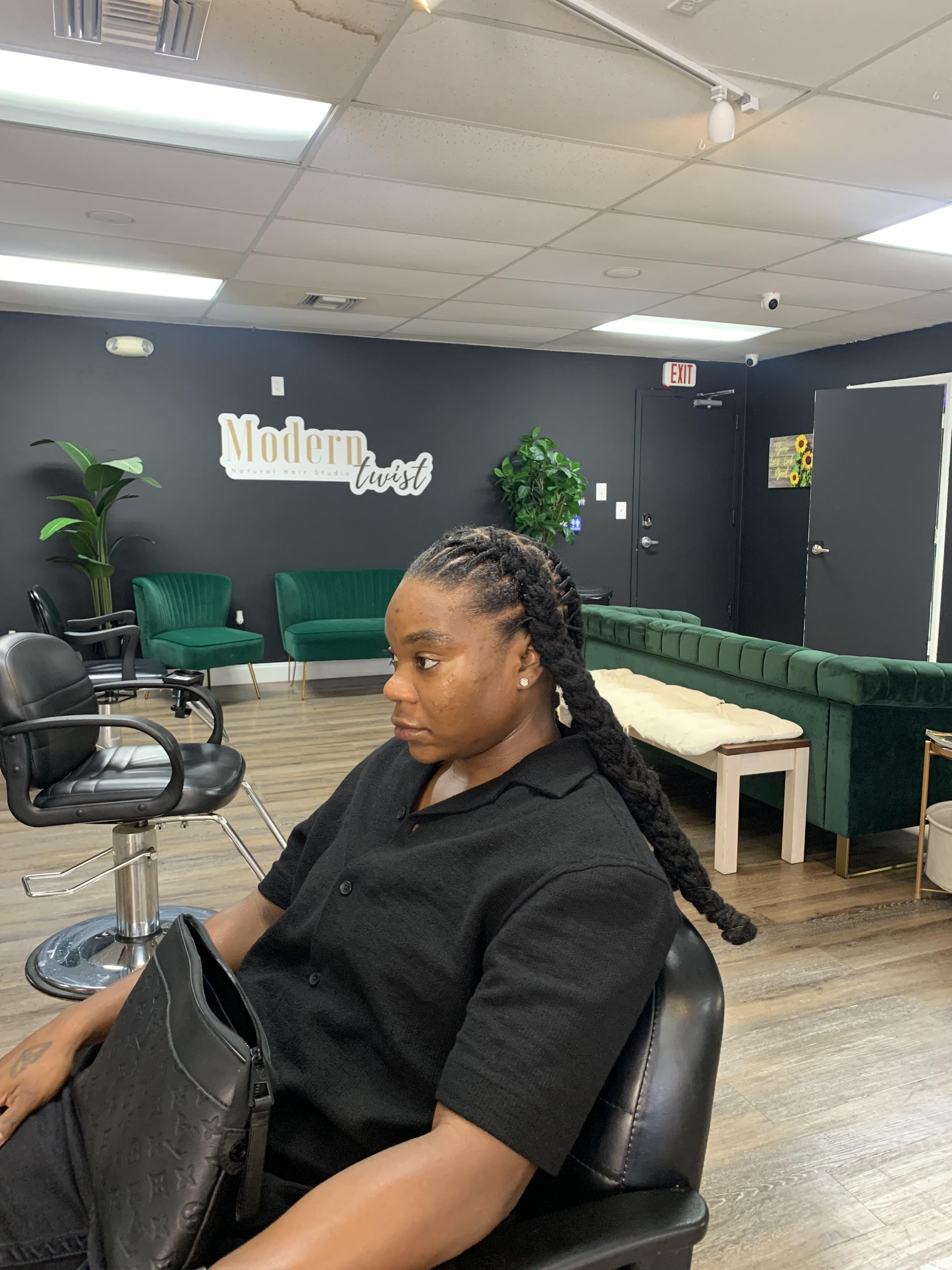 Black woman with long black braids and a hand tattoo sitting in a salon chair for a trim.