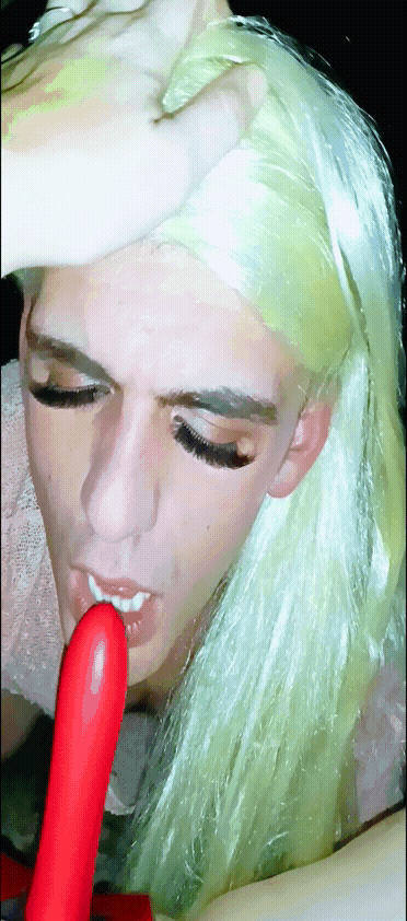 Sissy in a blonde wig performing a blowjob on a red dildo in a car with a woman