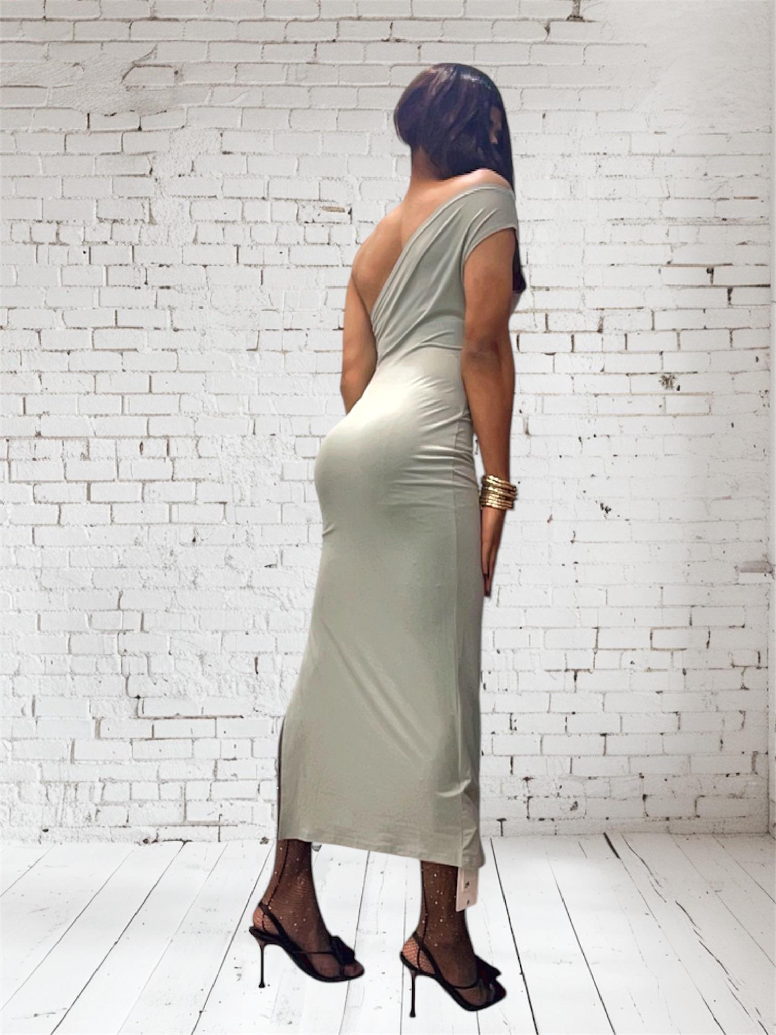 Desi sissy in a tight light green midi dress and black high heels posing against a white brick wall.