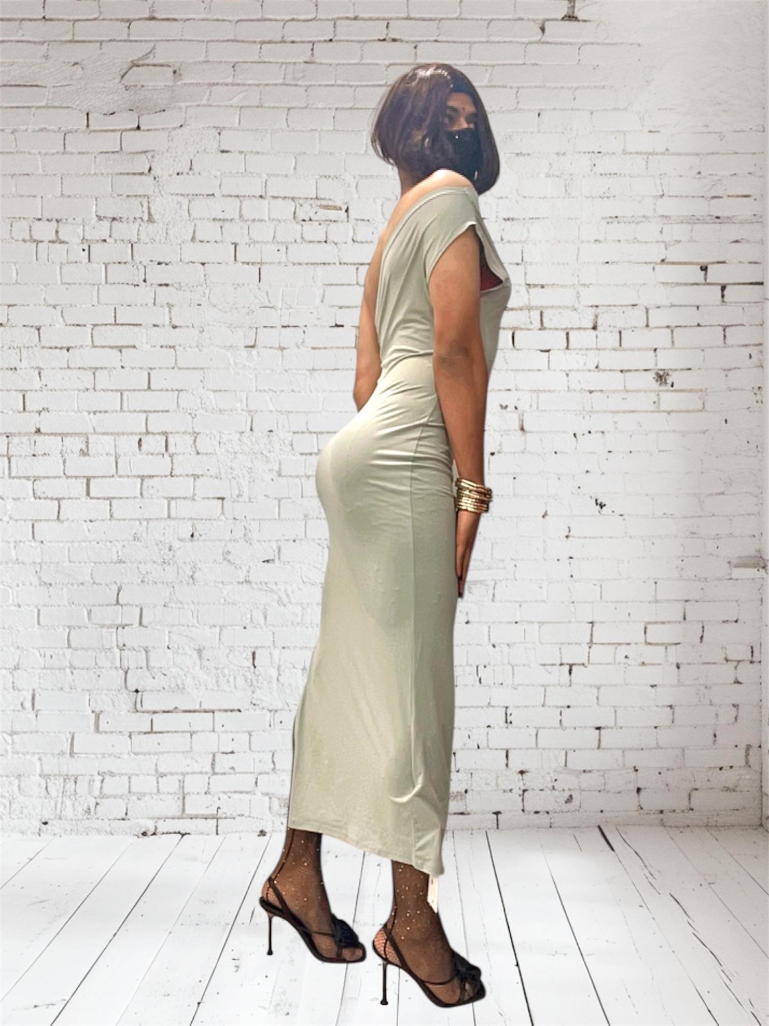 Desi sissy in a tight light green midi dress and black high heels posing against a white brick wall.