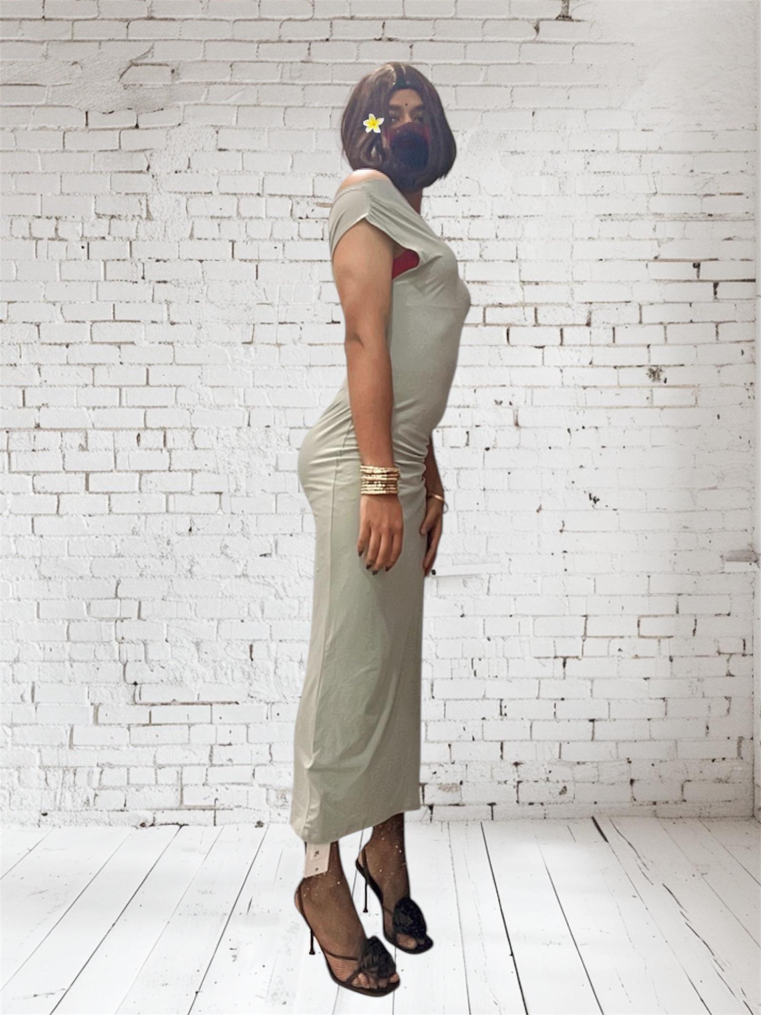 Desi sissy in a tight light green midi dress and black high heels posing against a white brick wall.