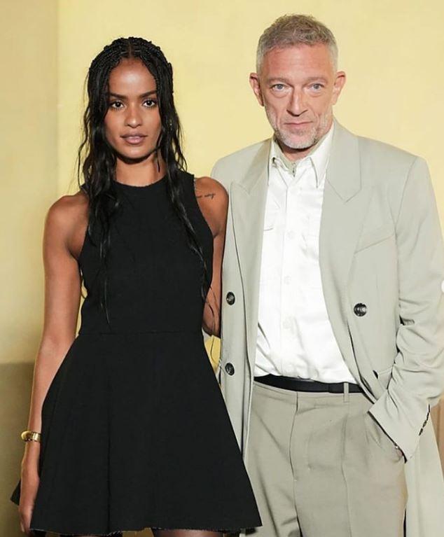 Narah Baptista in a black dress, white lace top, and grey suit, featuring her tattoos and tanned skin