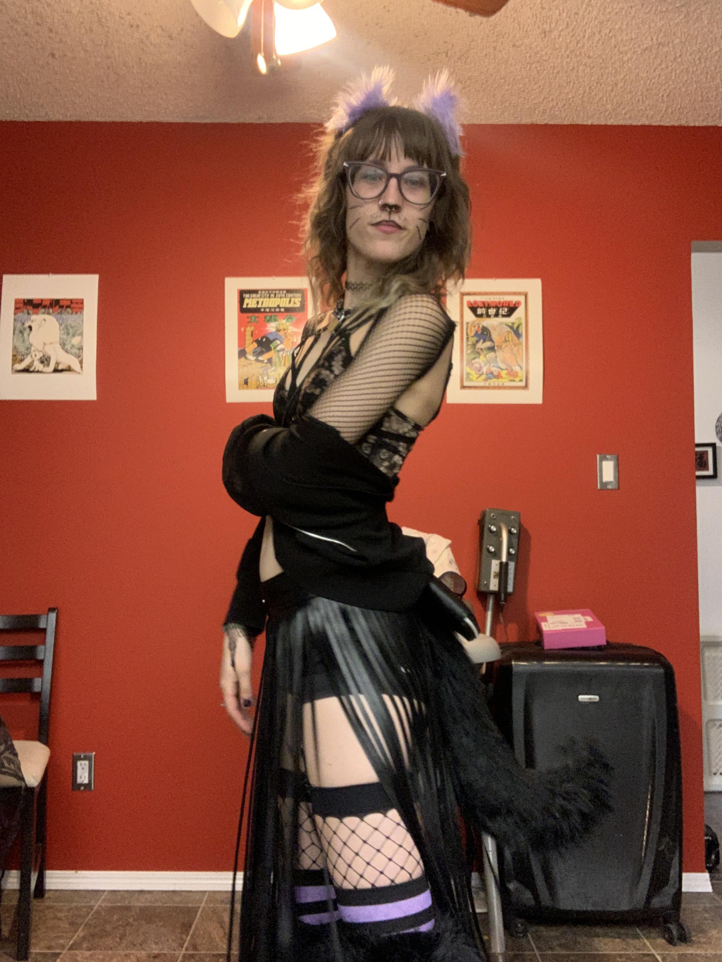 Blonde woman in a catgirl goth costume with a black fishnet top, skirt, and glasses posing for selfies