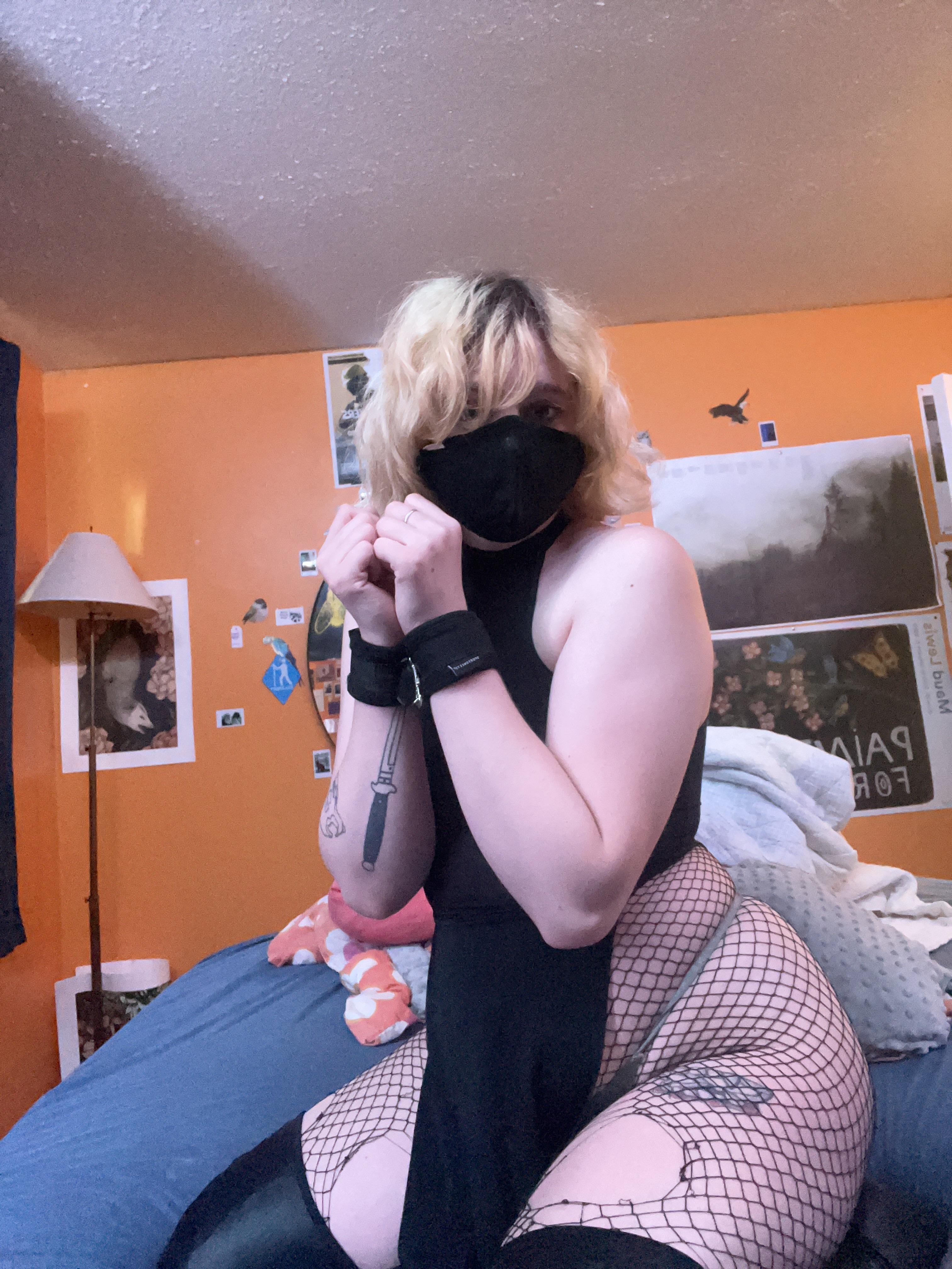 Blonde femboy in black lingerie, fishnets, and a red harness posing in a bedroom