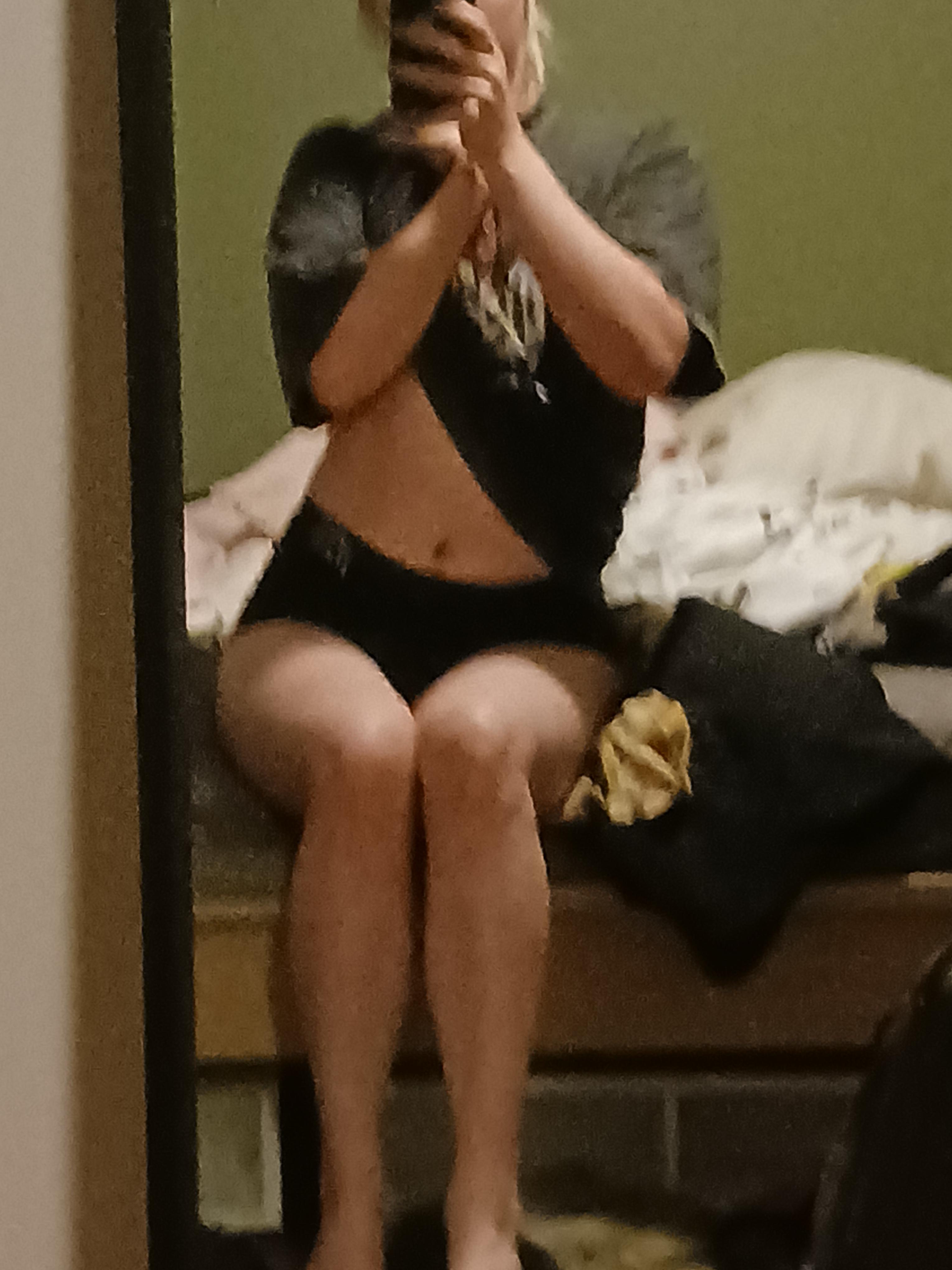 Curvy brunette femboy in black booty shorts posing on a bed, showing backside.