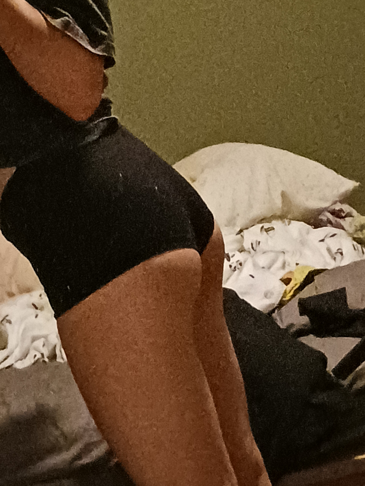 Curvy brunette femboy in black booty shorts posing on a bed, showing backside.