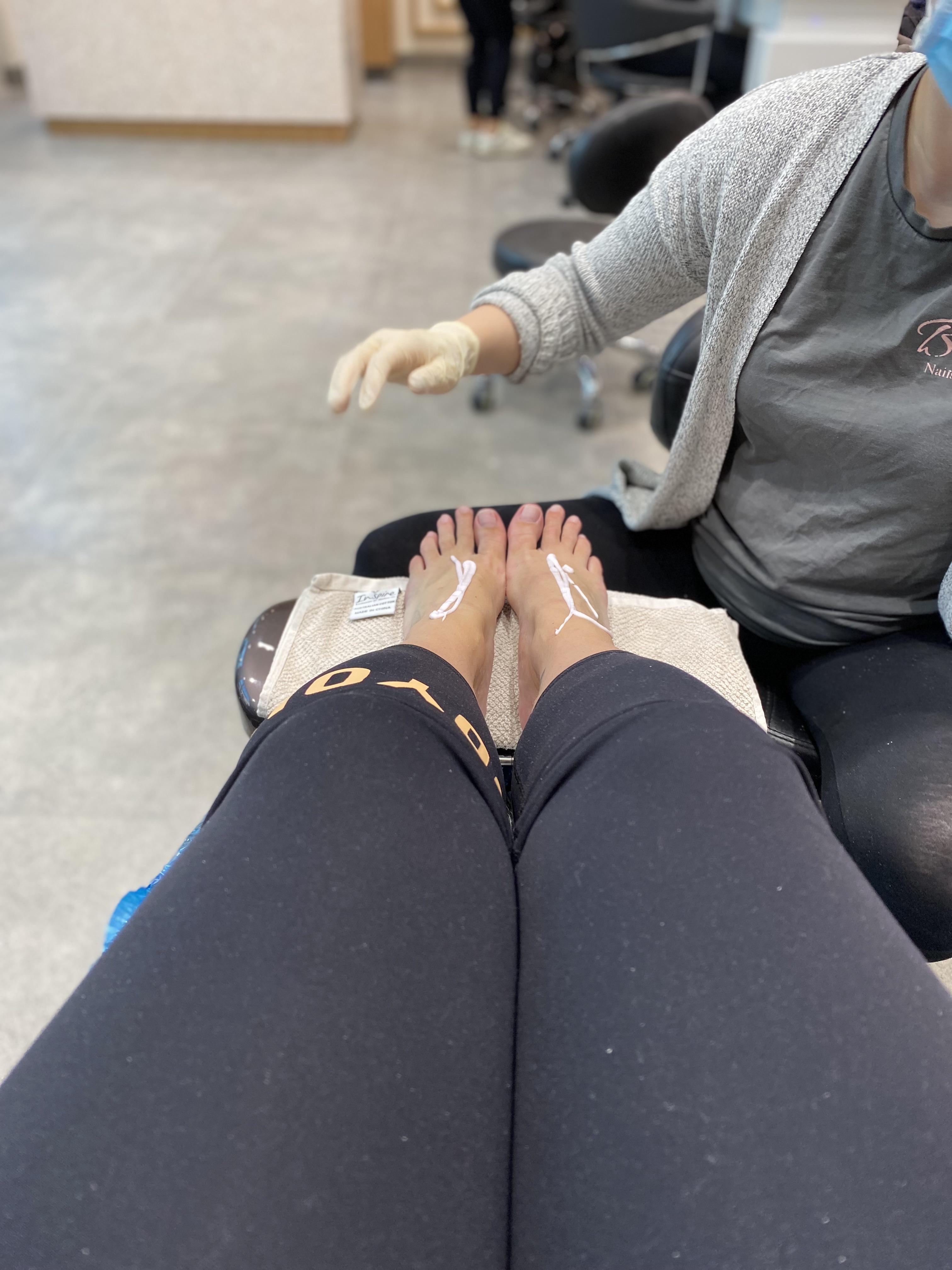Woman in black leggings receiving a pedicure, showing her bare feet, toes, and soles in a nail salon.