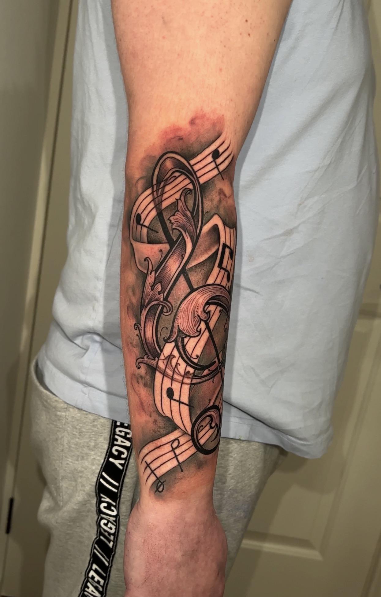 Musical-themed black and grey tattoo on a person's forearm with text reading LEGACY 1997