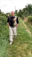 A heavy-set man in a black patterned shirt and white pants performing funny faces and meme actions in a village.