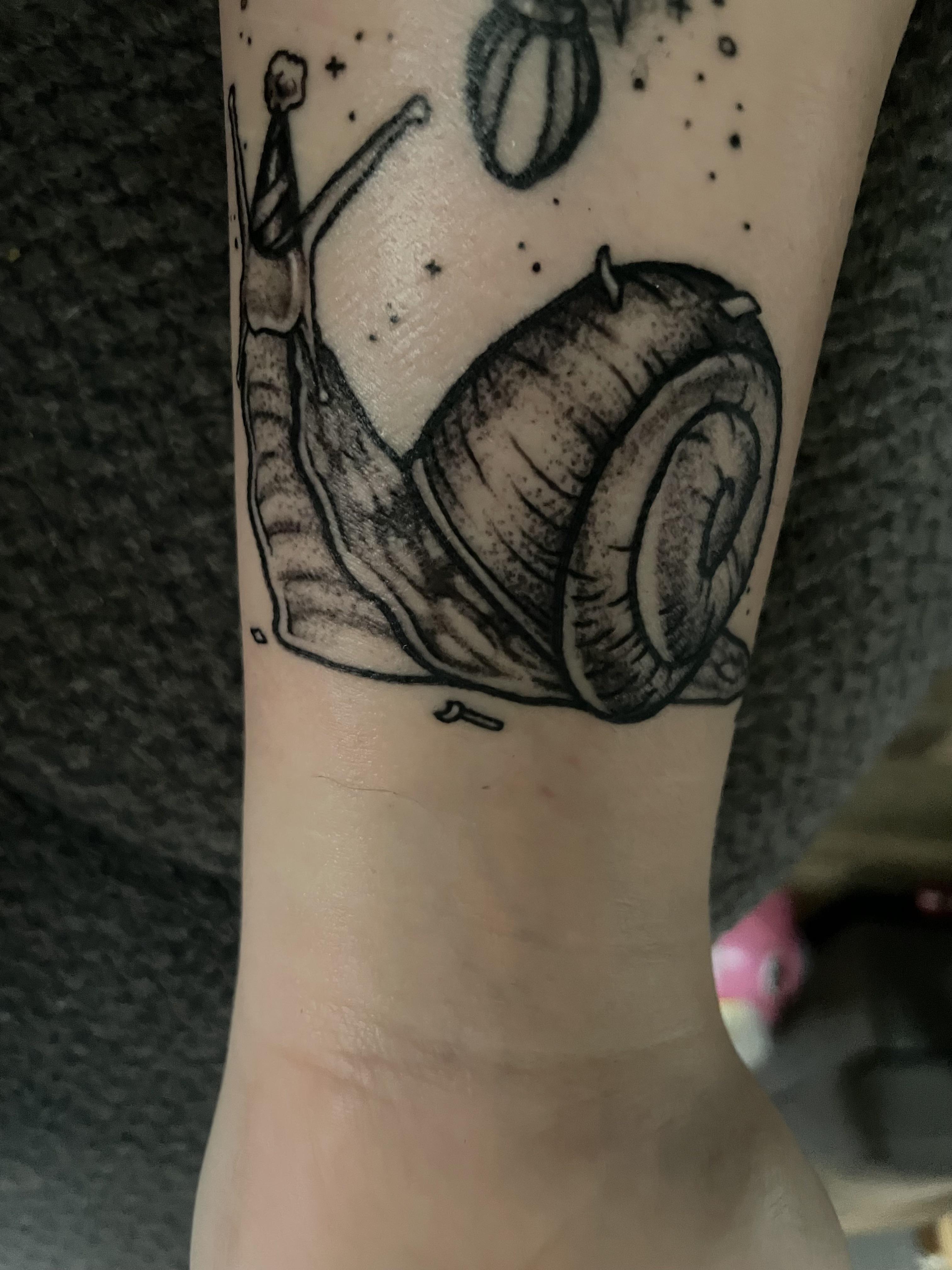 Fine line black ink tattoo of a kawaii snail with a party hat on a person's forearm