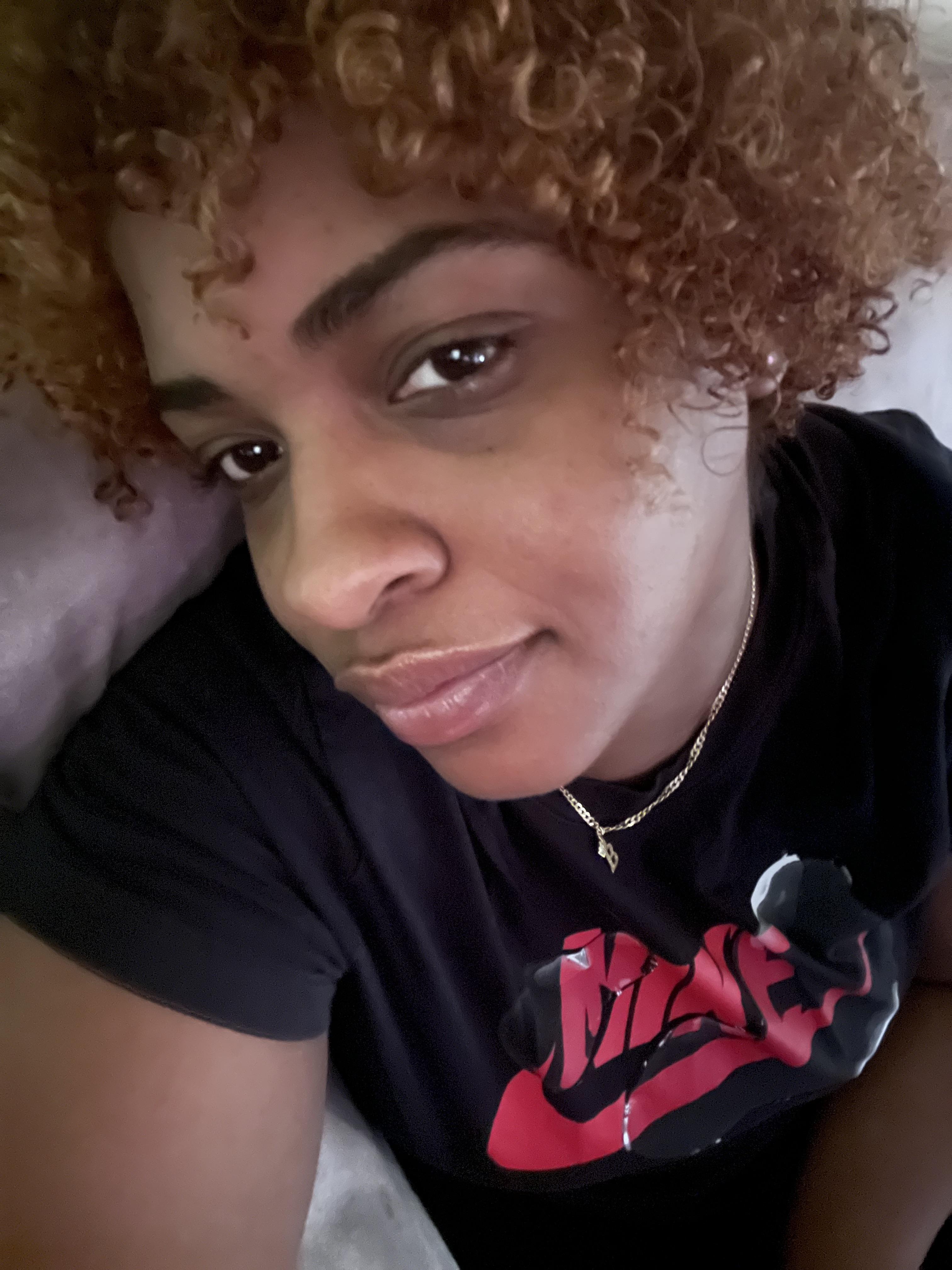 A person with curly blonde hair wearing a black Nike shirt and a gold chain necklace with a 'B' pendant.
