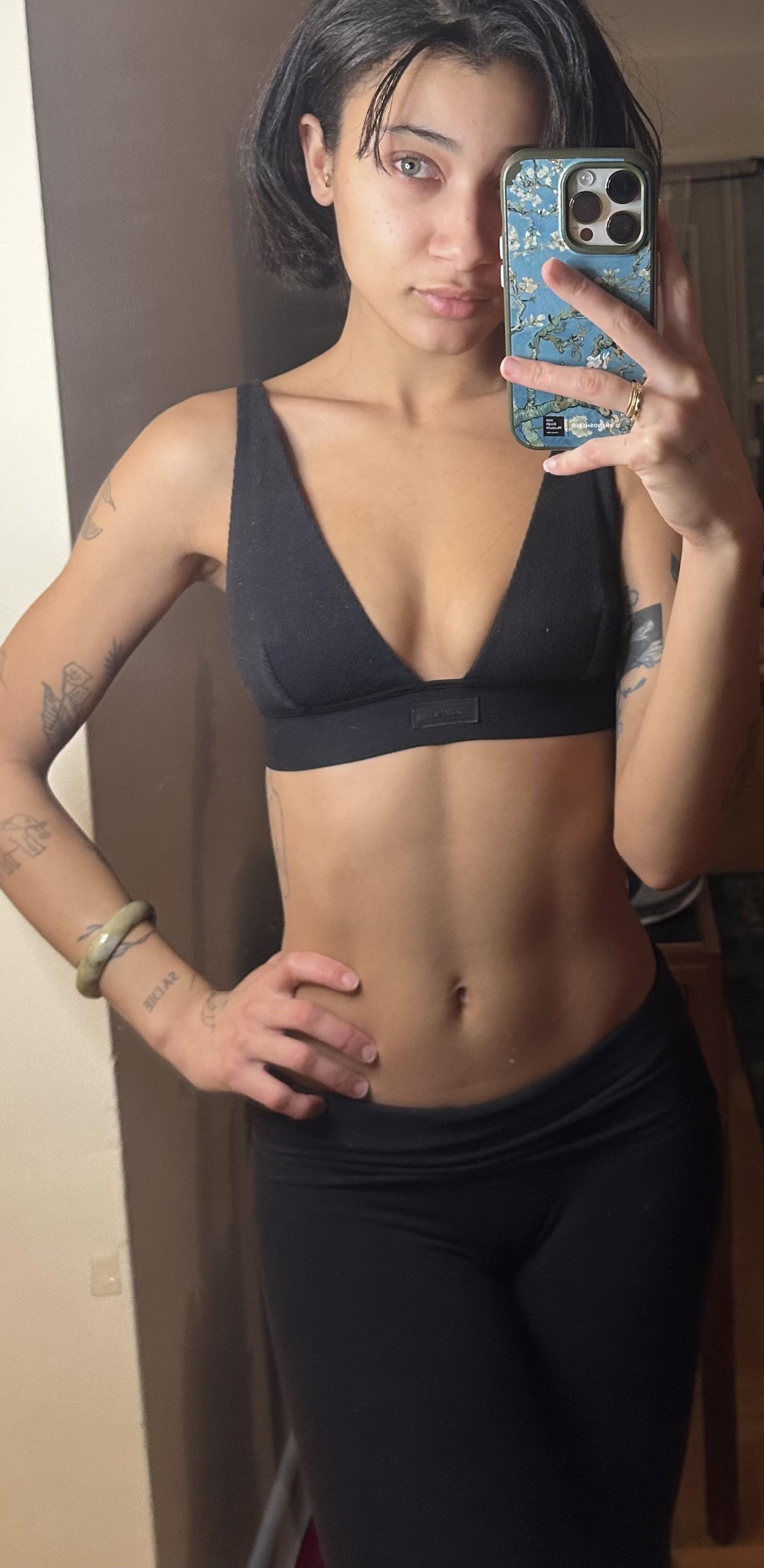 Mixed athletic woman in a black sports bra and leggings taking a mirror selfie, showing her midriff and tattoos.