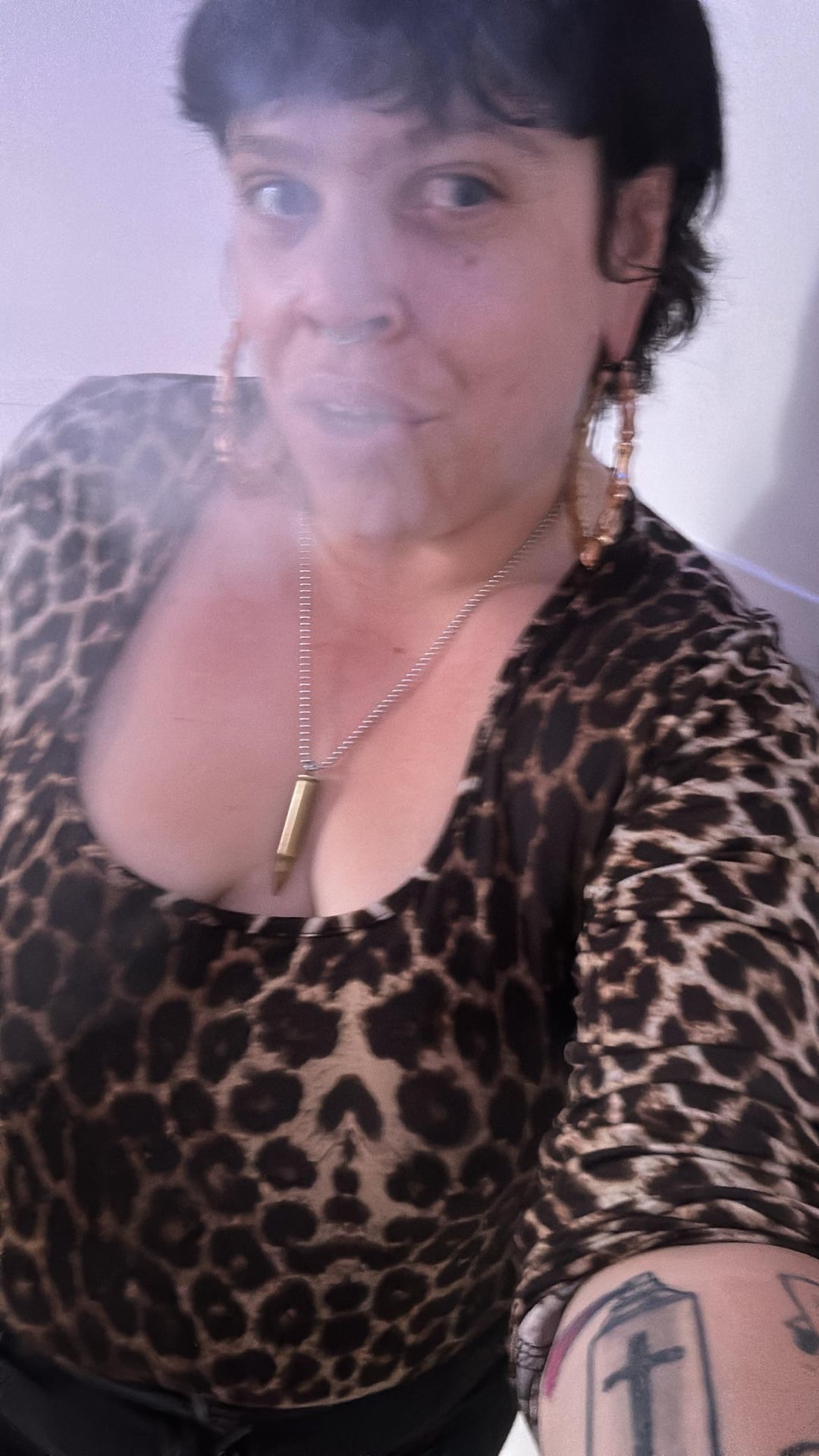A baddie with short dark hair wearing a leopard print v-neck top, gold hoop earrings, and a cross tattoo on her arm.