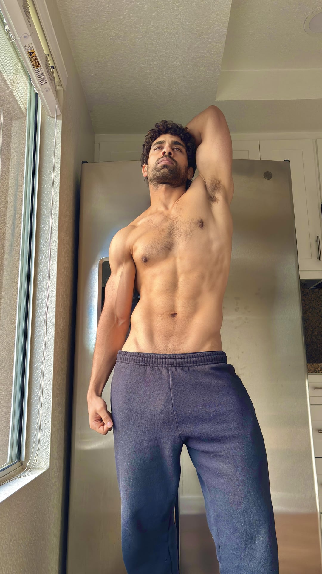 Muscular Indian man with curly hair posing shirtless and flexing in a kitchen selfie.