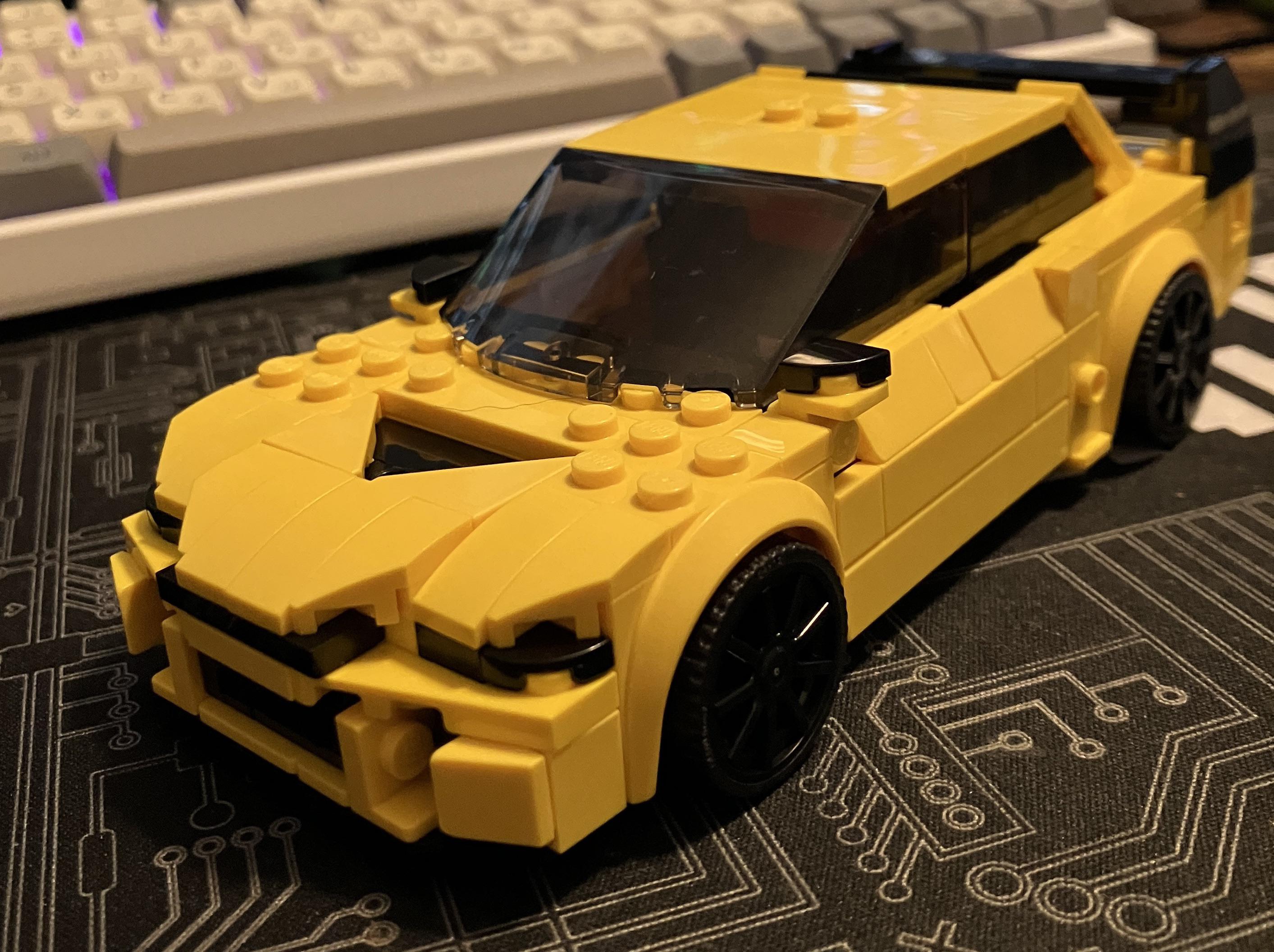 Yellow Lego Lancer sports car model on a gaming pc desk setup with a keyboard and desk mat
